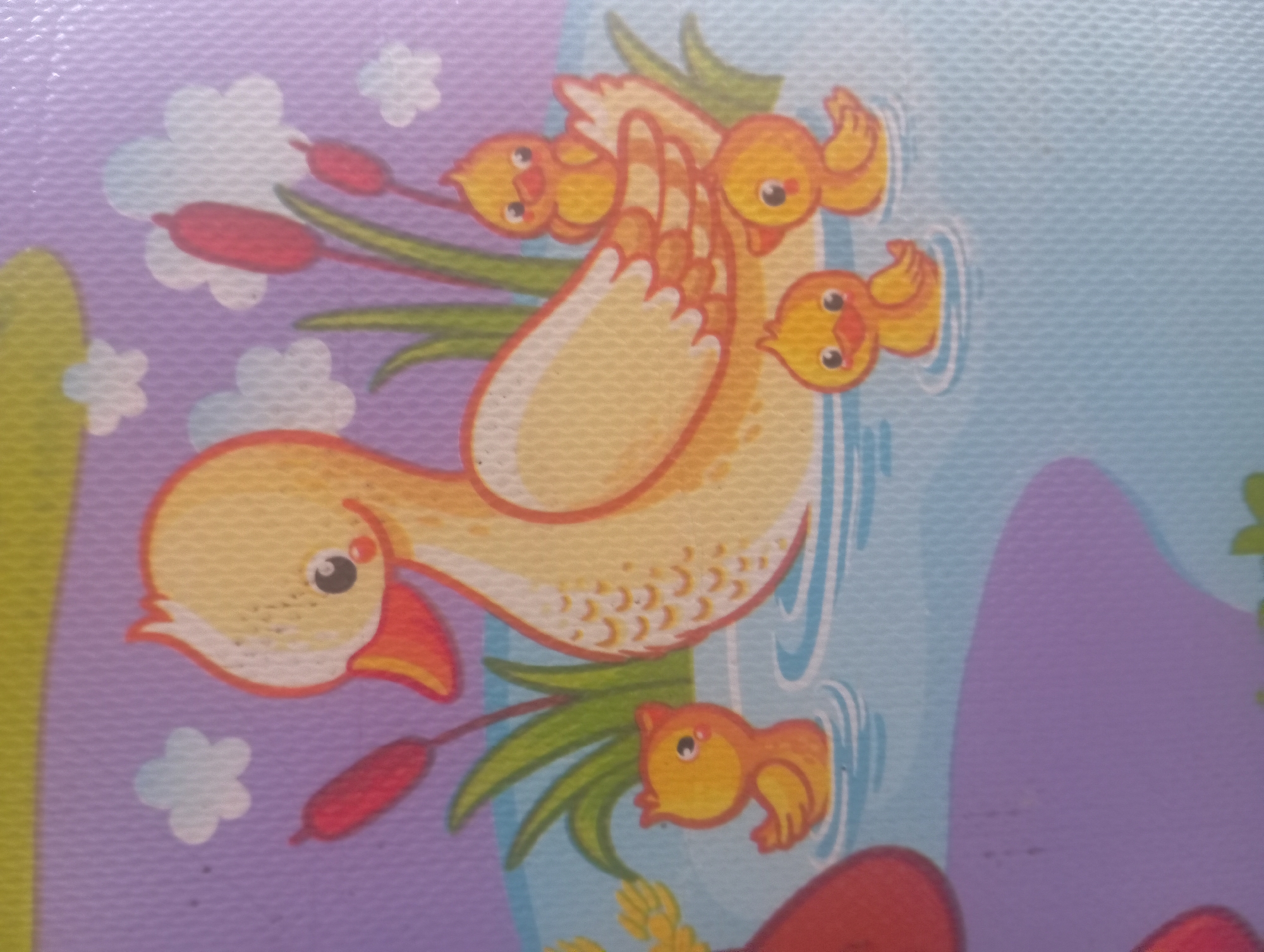 This is a close-up, overhead shot of a cartoon illustration depicting a mother duck and her ducklings in a body of water. The mother duck, larger and a pale yellow with orange accents on her beak and wings, is positioned centrally. Three small, bright yellow ducklings are around her. One duckling is to the lower left, another to the front right, and a third is positioned on the mother duck's back. The water is depicted with blue and white ripples. The background features stylized reeds with red tops, green leaves, and scattered white clouds against a purple sky. A large yellow and green shape is visible at the top edge of the frame. The image appears to be a detail from a children's mat or educational material, with a textured, woven background visible throughout. There is no visible indication of the city of Kutasari, Indonesia.