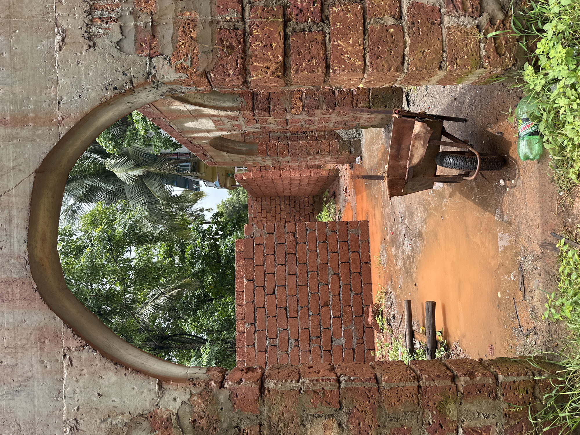 This image depicts an unfinished brick structure in Arambol, India. The scene is characterized by red brick walls, some still under construction, and a large arched opening in the foreground. The ground is muddy with a significant puddle of orange-tinted water. A rusty wheelbarrow stands on the wet ground, next to a discarded green plastic bottle. Beyond the immediate construction site, lush green trees and palm fronds are visible, with glimpses of a building and windows in the background, suggesting a location in or near a residential area. The lighting indicates it is daytime.