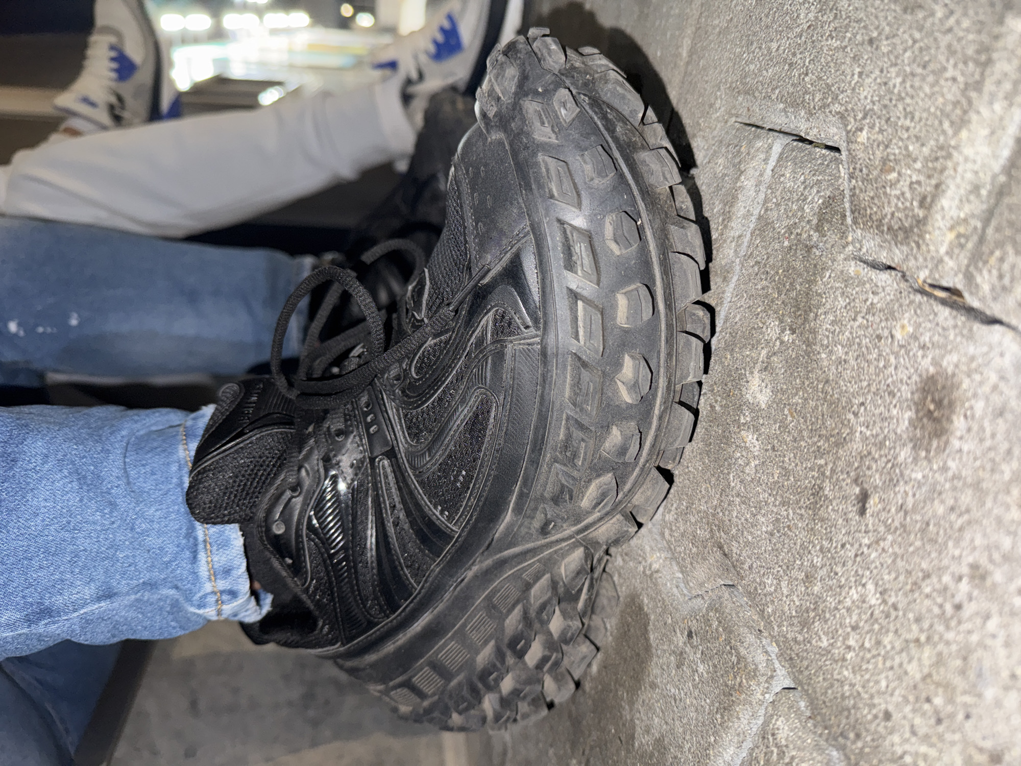 The image provides an angled, close-up view focusing on a person's legs and footwear, set against a textured concrete surface. The main subject is wearing light blue jeans, cuffed at the ankles, and a black athletic shoe. This shoe features a thick, rugged sole with a distinctive, deep lug pattern, suggesting durability and traction. The upper part of the shoe appears to be constructed from a combination of mesh and synthetic materials, with black laces tied.
The black shoe is positioned with its sole resting on a light gray, granular concrete or stone surface, which shows signs of wear, including visible cracks and irregularities. In the blurred background, another person's lower legs and feet are visible. This individual is wearing white pants and white sneakers with blue accents. The scene appears to be illuminated by artificial lighting, creating a soft glow in the background.
The setting suggests an indoor or covered public space, possibly a stadium, arena, or a large building with concrete flooring, consistent with the location context of Sevlievo, Bulgaria. The activity depicted is casual, with the individuals seemingly sitting or resting. No visible text is present on any objects or in the environment.