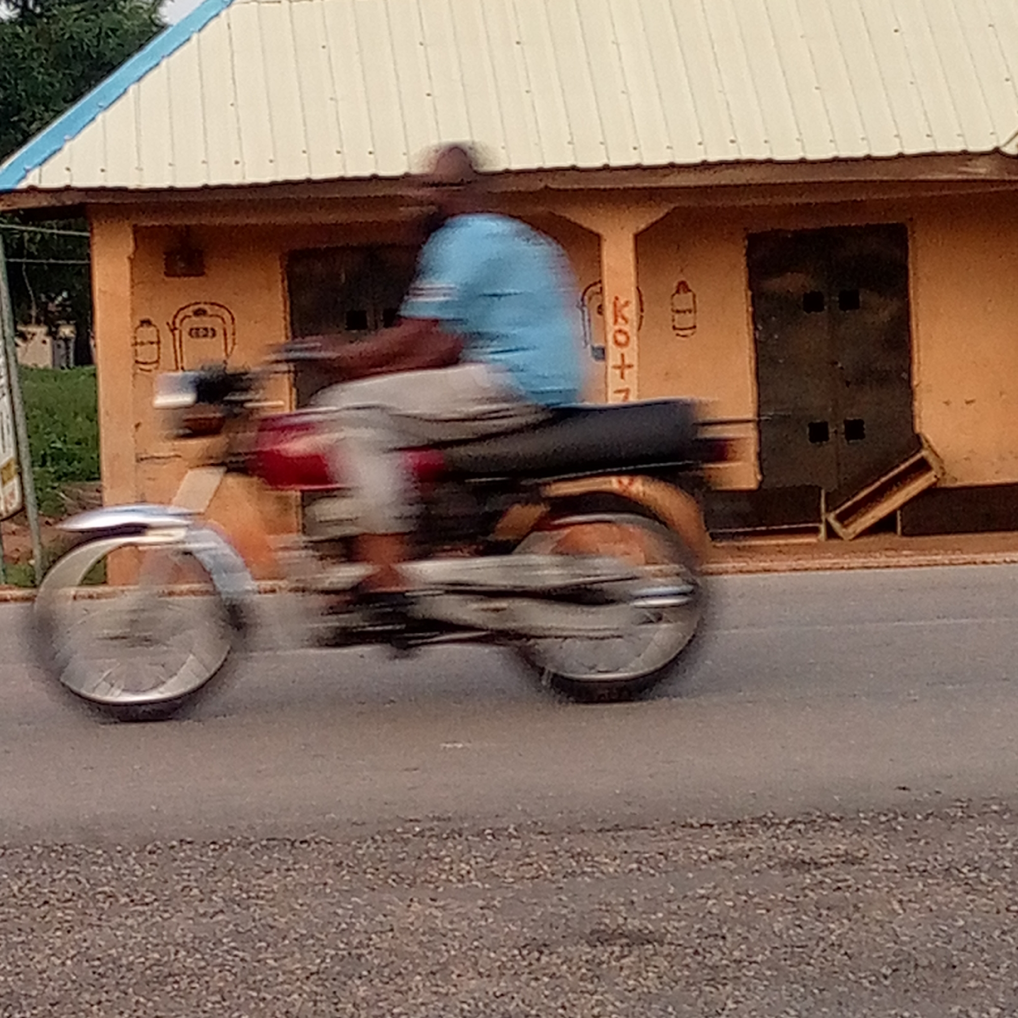 The image captures a person riding a motorcycle along a paved road in Mararaba, Nigeria. The main subject, an adult wearing a light blue t-shirt and light-colored shorts, is in motion on a red and black motorcycle, both appearing significantly blurred, especially the wheels, due to a panning photographic technique that highlights movement against a relatively stable background.
In the background stands a single-story building with a tan or light orange facade and a white corrugated metal roof, distinguished by a blue trim along one edge. The building features dark doors, one of which is visible on the right. On the building's wall, several distinct brown line drawings are visible. To the left, two cylinder-like objects are drawn, one containing the text "(E23)". Further to the right, another cylinder drawing and partially visible text "KO + T" can be seen. Green foliage is visible behind the building on the far left. The scene is illuminated by daylight, and the road surface shows a coarse texture. The image conveys an everyday street scene with a focus on motion.