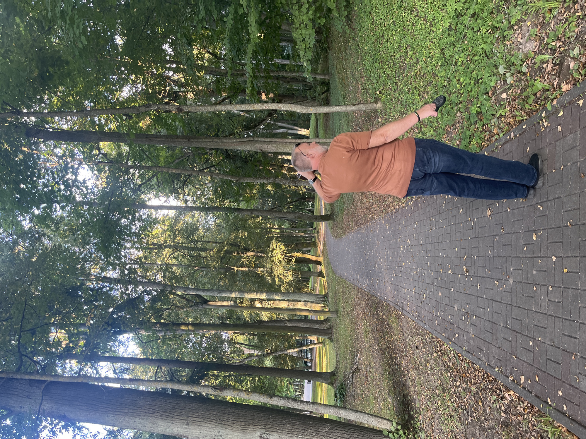 An adult male wearing a brown t-shirt and blue jeans stands on a paved pathway in a park. He is facing away from the camera, towards the left side of the image, with his right hand touching the back of his head. He holds a black leash in his right hand, extending towards the right side of the image. The pathway is made of gray pavers and is bordered by grass and fallen leaves. Tall trees with green foliage line the pathway, creating a shaded environment with dappled sunlight filtering through the leaves. In the background, more trees and greenery are visible, along with what appears to be park furniture. The scene is set in a park in the city of Lahojsk, Belarus.