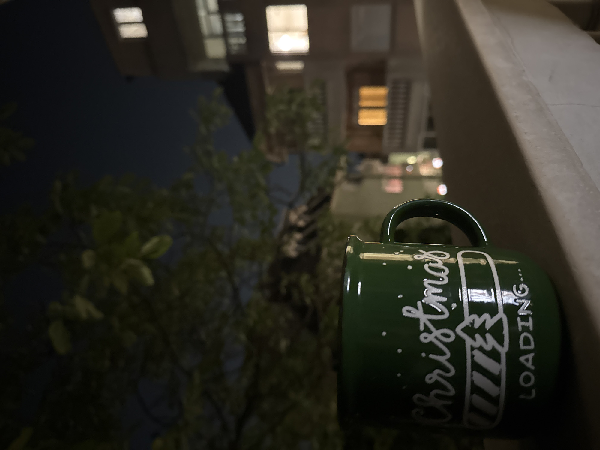 A green mug with the words "Christmas...LOADING" written on it sits on a ledge in what appears to be a city. The mug is positioned in the foreground, with the city lights and building structures blurred in the background. There's also the presence of green foliage, suggesting a natural element within the urban setting. Given the presence of the word "Christmas" on the mug, it seems to be a festive scene. The photo does not contain enough information to determine the precise location.