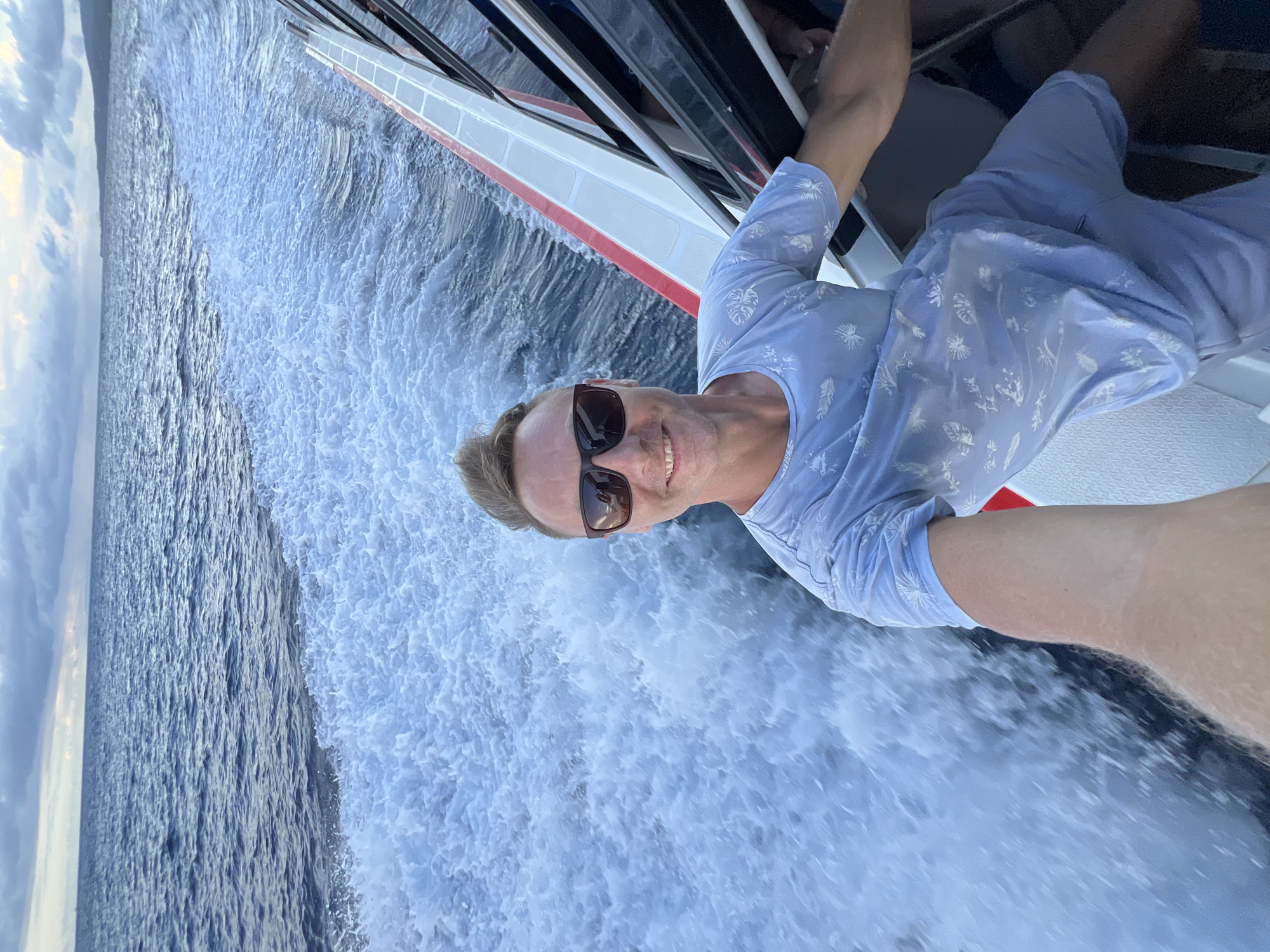 This image is a selfie taken by a smiling man on a moving boat, likely off the coast of Nusa Penida, Indonesia. The man, with fair skin and blondish hair, wears dark sunglasses and a light blue short-sleeved shirt adorned with white patterns resembling leaves or abstract shapes, paired with light blue shorts. He is positioned on the boat, holding the camera to capture himself and the dynamic maritime scene.
The boat, partially visible with its white hull and a red stripe, is actively traversing the water. A significant trail of white, churning wake is created by its movement, contrasting with the darker, choppy blue of the open ocean. In the background, the sea extends to a distant horizon beneath a cloudy, light blue sky, where a faint silhouette of land or an island can be seen.
The man's broad smile suggests a feeling of happiness and enjoyment during the boat trip. The scene conveys a sense of adventure or leisurely travel on the water, likely during the daytime under bright, albeit cloudy, conditions. No discernible text is visible within the image.