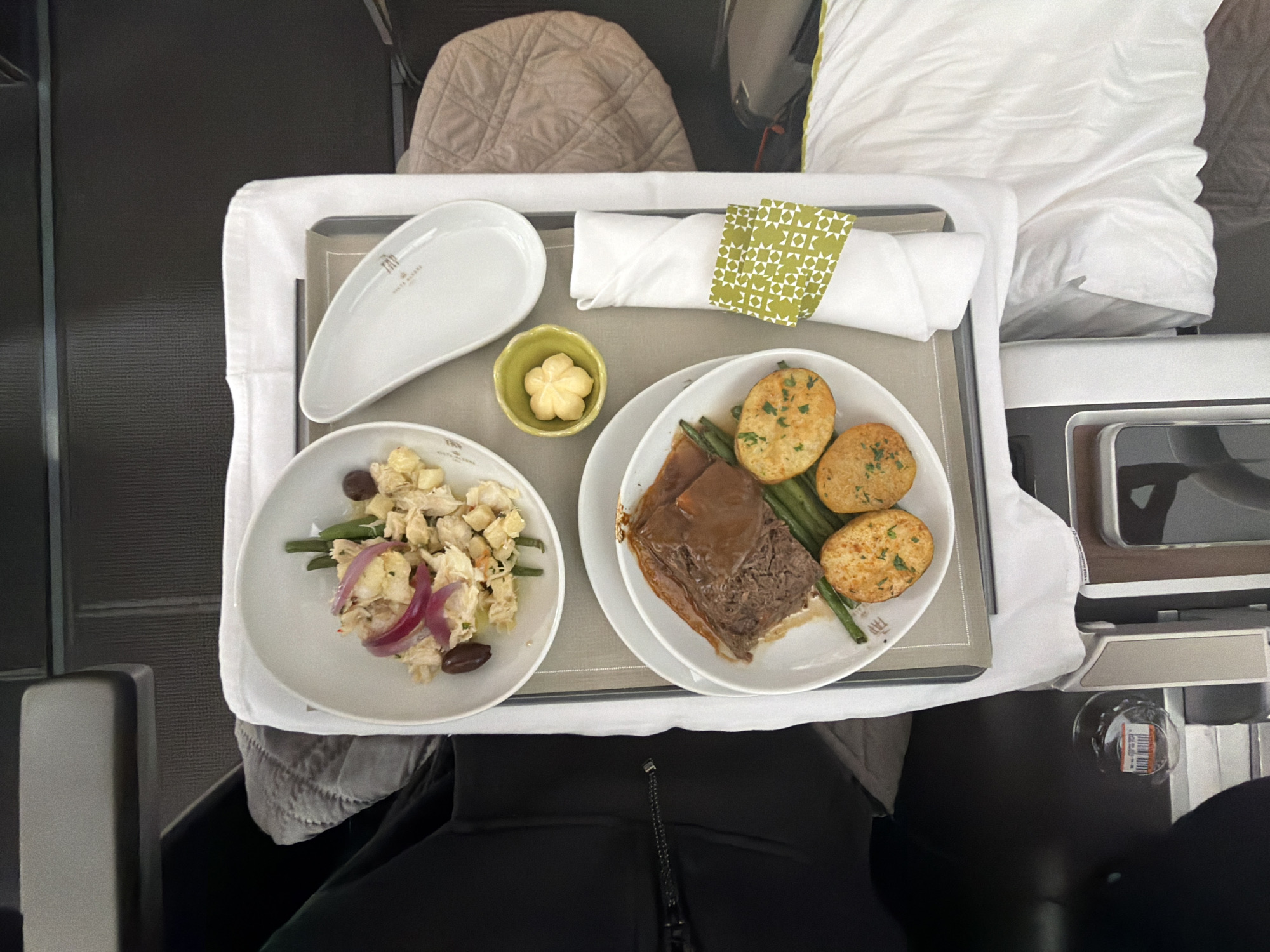 An overhead view captures an inflight meal served on a tray table within an airplane cabin, likely in transit to or from Mississauga, Canada. The meal consists of two primary dishes on white ceramic plates: an appetizer salad featuring shredded protein, red onion, green beans, and black olives, and a main course of sliced braised meat with gravy, accompanied by green beans and three pan-fried potato patties. A small green bowl holds a flower-shaped pat of butter. Tableware includes a white linen napkin with a green and white geometric patterned ring, a gray placemat, and a distinctive elongated white dish bearing the "TAP PORTUGAL" and "WORLD CLASS" logos. The tray is positioned on a dark-colored seat-back table. The immediate environment shows portions of dark fabric seat upholstery, a metallic armrest on the left, a quilted beige cushion on an adjacent seat to the right, and a white sheet or blanket in the lower right. A small, clear water bottle rests on a side console in the lower left. Only a segment of dark clothing from an unseen individual is visible on the left.