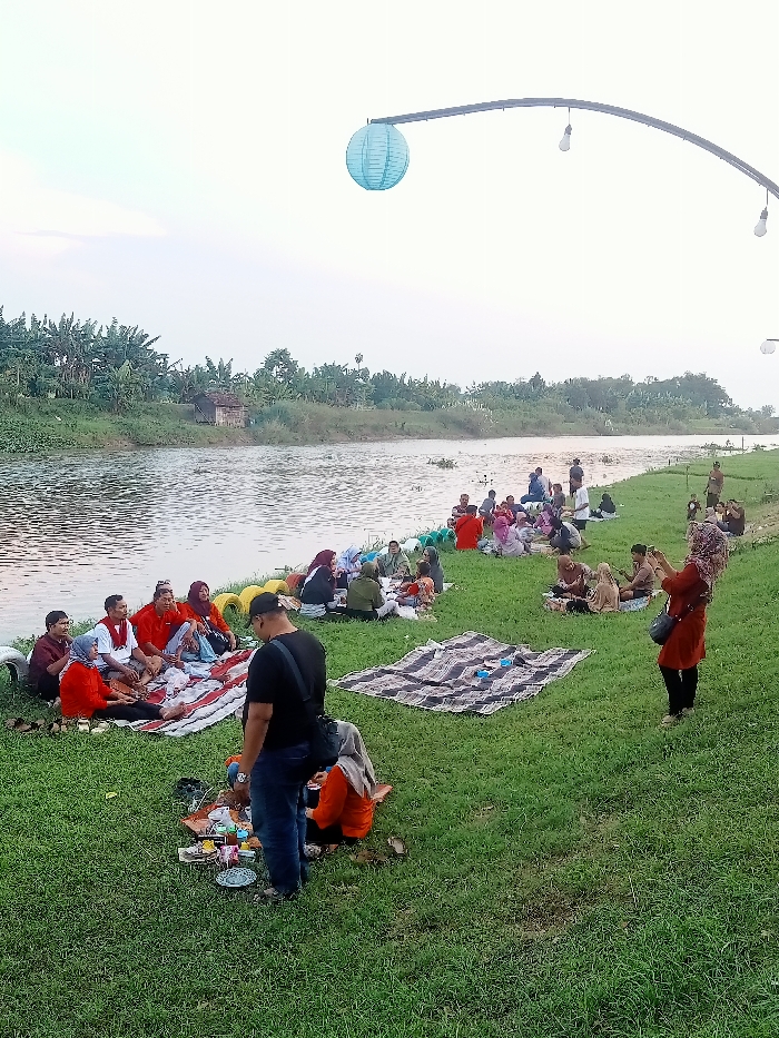 The image shows a group of people picnicking on the grassy bank of a river in Jakenan, Indonesia, located at coordinates -6.758 latitude and 111.075 longitude. The individuals are sitting on blankets spread out on the ground, some are eating and others are talking. There are several objects scattered on the ground, including food containers, plastic cups, and a blue and white striped blanket. One woman is standing and using her phone. There is a blue paper lantern hanging from an overhead light fixture in the upper left side of the image. In the background, there is a river with a lush green bank and several trees. The sky is overcast. The scene is peaceful and relaxed.