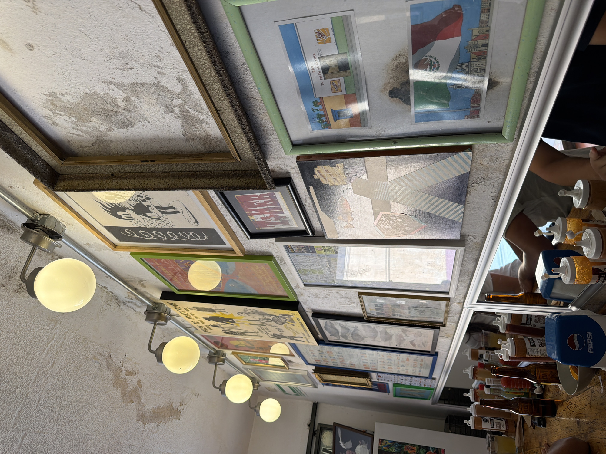 This image depicts the interior of a casual establishment, likely a bar or restaurant, located in San Andrés Cholula, Mexico. The textured, off-white walls are extensively covered with an eclectic array of framed artworks, posters, and prints. Notable wall decorations include a "FIESTA DE MONSTRUOS" poster, an illustration of a black cartoon character, and a light green-framed print containing two smaller images: one of a building labeled "BAR EL SALÓN DE LA YAMA" with a Sol beer logo, and another featuring a Mexican flag over a city skyline with a prominent church dome, consistent with Cholula's architecture. An empty, ornate gold-toned frame is positioned high on the right wall. Five spherical, frosted glass light fixtures are mounted on exposed metallic conduit along the ceiling, providing illumination. Below the wall art, a long mirror reflects a wooden tabletop. This surface is populated with numerous condiment squeeze bottles, several amber glass beer bottles, a white bowl filled with orange citrus, and a blue Pepsi-branded napkin dispenser. Partial reflections in the mirror reveal the arms and torsos of at least two individuals, indicating their presence at the table. The walls and ceiling exhibit visible textural variations and areas of discoloration.