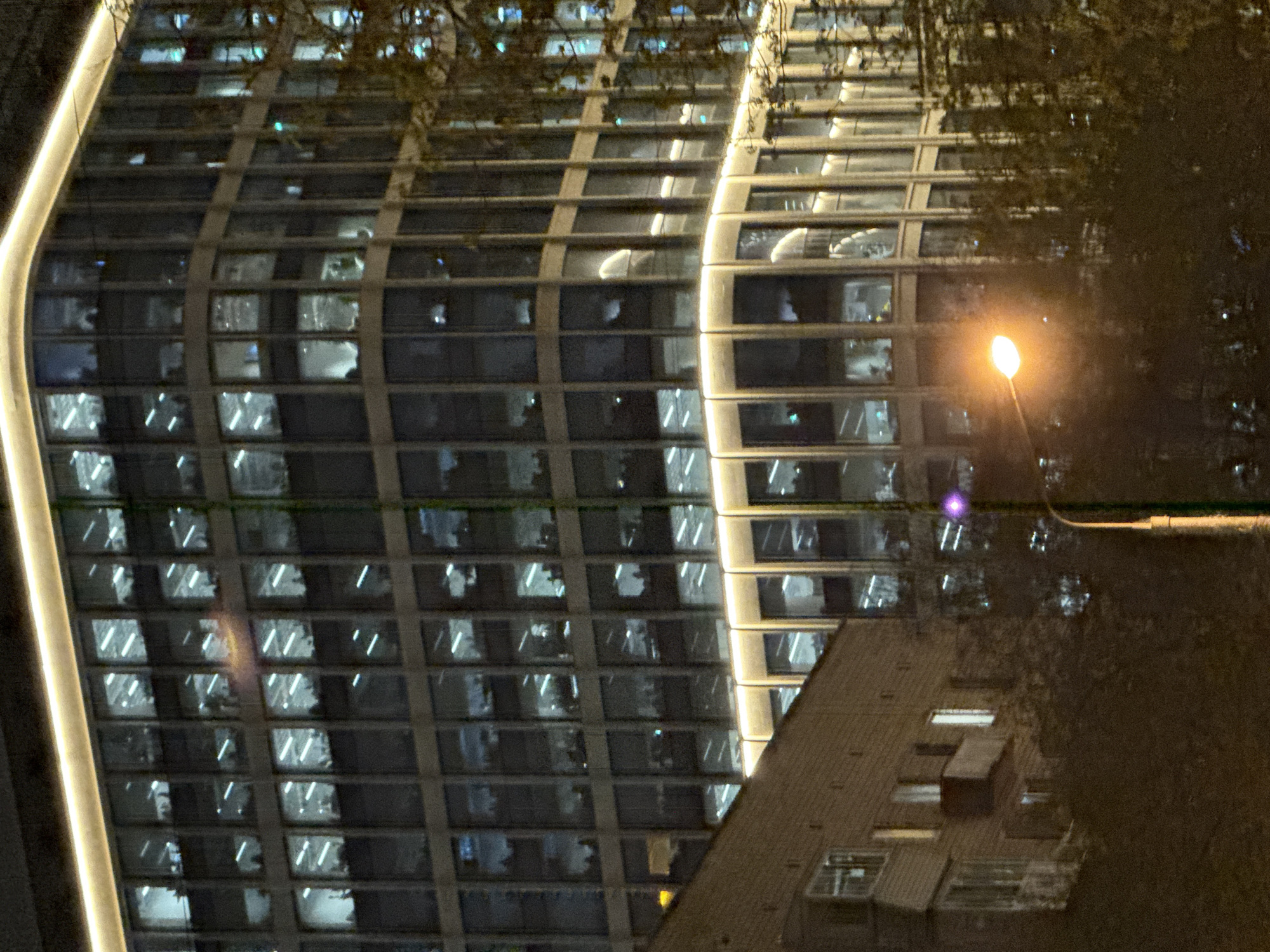 At night, a curved modern building with many windows is illuminated with lights. A street lamp provides additional illumination. The scene appears to be in Moscow, Russia. The building is partially obscured by a tree. Another, smaller building is also partially visible.