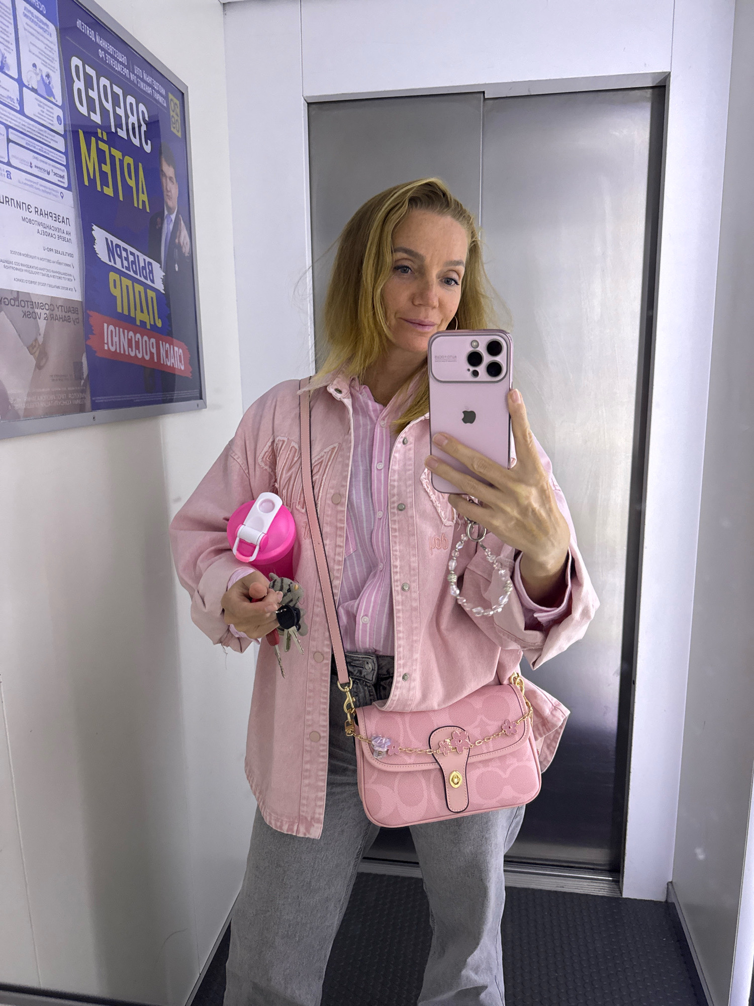 A woman is taking a selfie in what appears to be an elevator, with a poster on the left wall. She is wearing a pink denim jacket over a pink striped shirt and gray jeans. She is holding a pink water bottle in her left hand and her cell phone in her right hand, taking the picture. A pink shoulder bag with a gold chain and floral embellishments is visible on her right side. The poster on the wall is in Russian and features a man's face. The environment suggests an indoor public space, possibly a residential building or commercial establishment in Sinyavino, Russia, given the presence of a Cyrillic-language poster.
