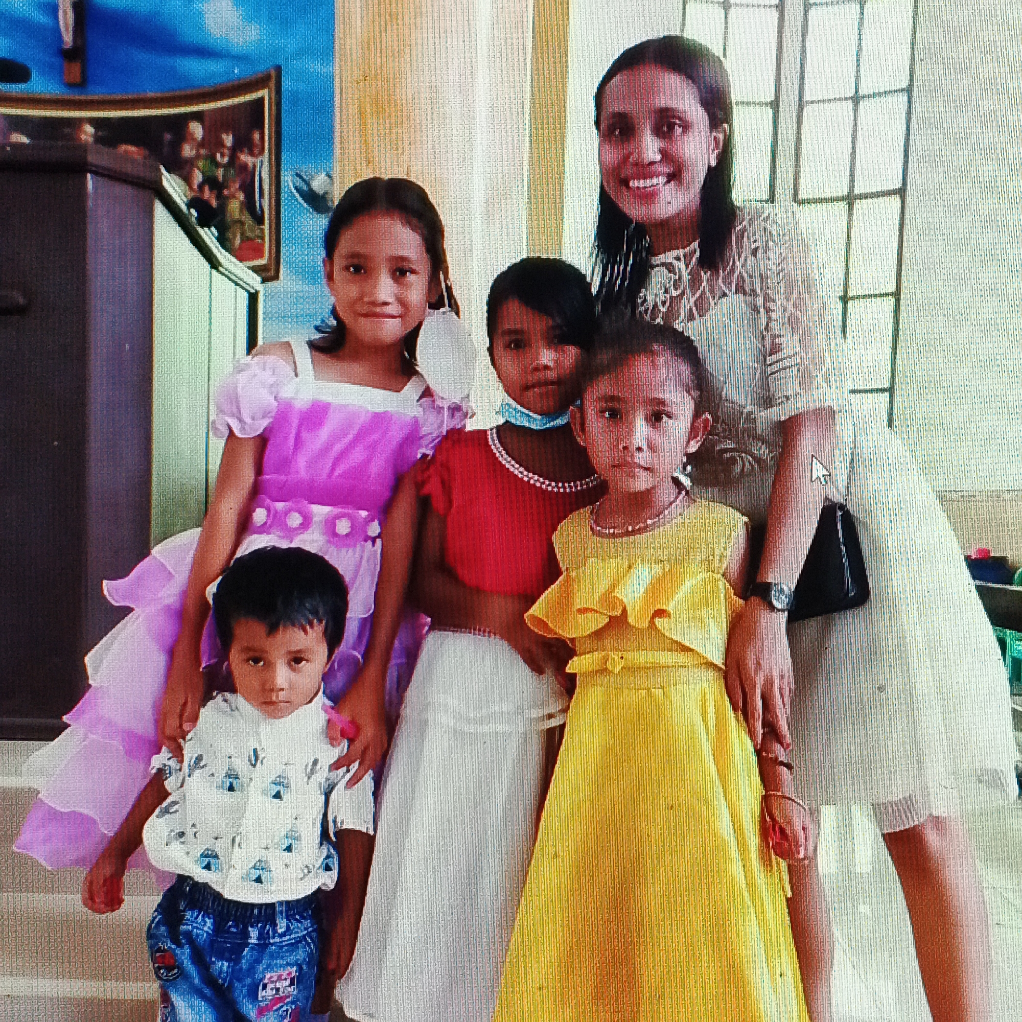 The image depicts a group of five individuals, one adult woman and four young girls, posing for a photograph indoors. The woman, who appears to be East Asian with dark hair, is smiling and wearing a white lace dress. She is standing behind the girls. The girls are dressed in colorful attire. The leftmost girl wears a lilac dress, the middle girl in the front wears a red top and a white skirt, and the rightmost girl wears a yellow dress. A young boy in a patterned shirt and blue jeans is standing in the front left. The background appears to be a room with a patterned wall or curtain, and to the left, there is a dark wooden cabinet with a framed painting. The scene suggests a celebratory or formal occasion, possibly a family gathering or a church event, given the painting which might be religious. The lighting suggests an indoor setting, with the overall atmosphere appearing warm and familial. The provided crops confirm the details of the individuals' clothing, facial expressions, and some background elements, including a possible window frame behind the woman. The question about the location in Ende, Indonesia cannot be answered from the image alone; it requires external information not present in the visual data.