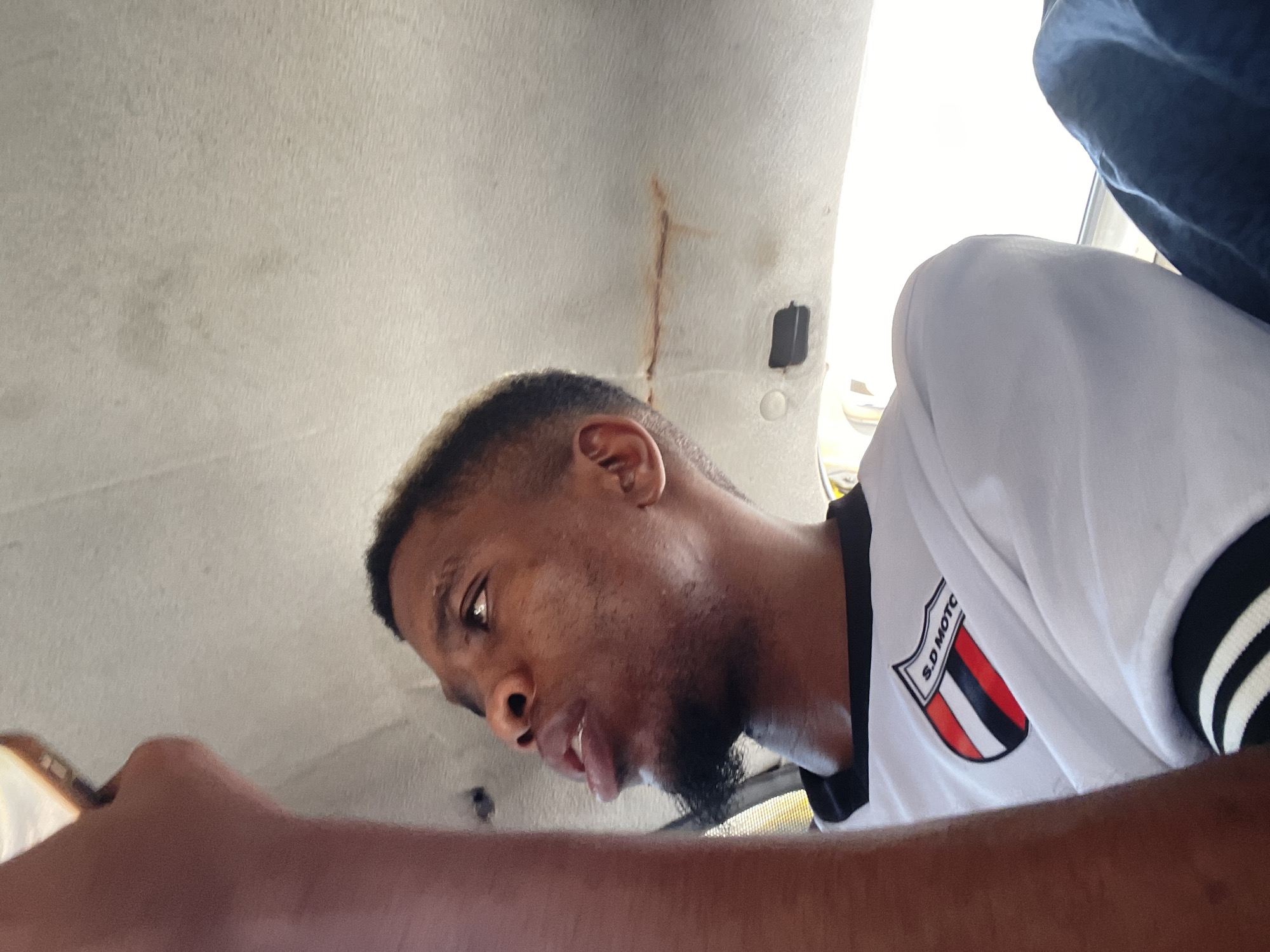 The image shows a person with a beard and a short haircut, wearing a white shirt with black trim and a patch with "S.D MOTO" on it. They are looking down, possibly at a mobile phone in their hand. The background shows the interior of a vehicle, specifically the ceiling which is stained. The location is potentially Lagos, Nigeria.