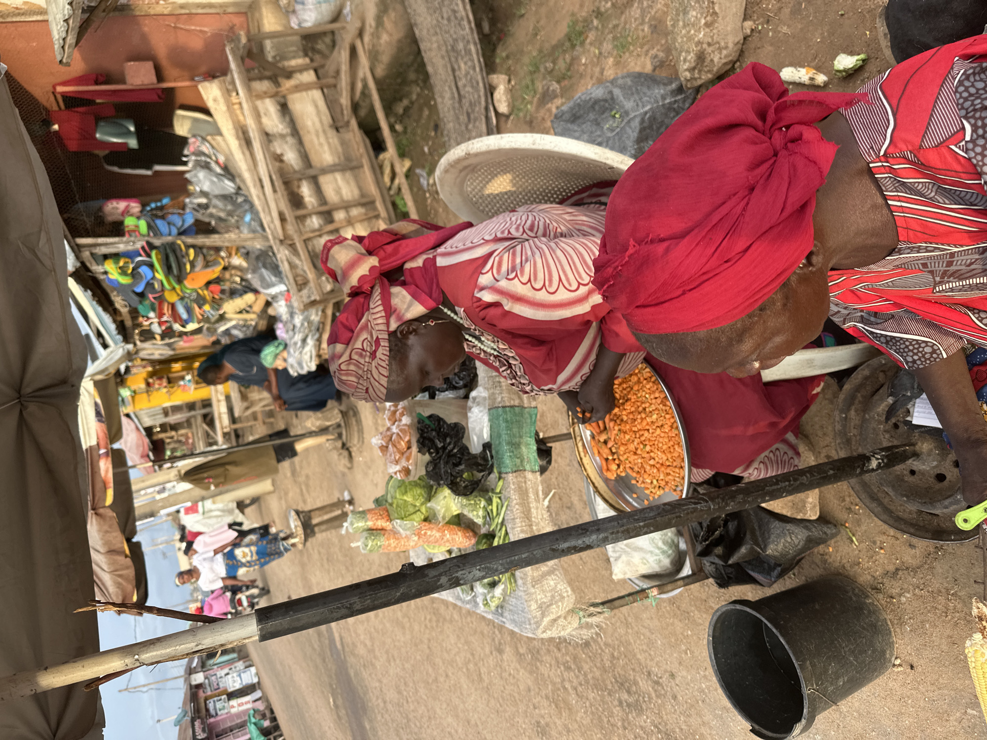 In the city of Narayi, Nigeria, a market scene unfolds under a partially unfurled beige awning. The foreground is dominated by two women, both wearing vibrantly colored headscarves and traditional African attire. One woman, seated on a white plastic chair, is actively engaged in preparing food, her hands manipulating chopped orange vegetables on a metal tray. Her red and white patterned robe is partially visible. Next to her, a large black bucket sits on the dusty ground, and behind her, a metal pole with a circular base supports the awning.
A table is set up behind the seated woman, laden with produce. Bundles of carrots, some wrapped in plastic, are prominent, alongside heads of cabbage and other green vegetables. Bags of produce and a green cloth are also visible on the table. The second woman, in the lower right of the frame, is also wearing a red headscarf and a red, white, and black striped outfit. She is leaning forward, looking down, with her hands near the chopped vegetables.
The background reveals the bustling market environment. Several people are walking in the distance along a dirt road, some on foot and one on a motorcycle. Stalls with various goods, including colorful flip-flops and clothing, are set up along the road, with wooden structures and corrugated metal roofing. The overall impression is a lively, everyday market in a Nigerian urban setting.