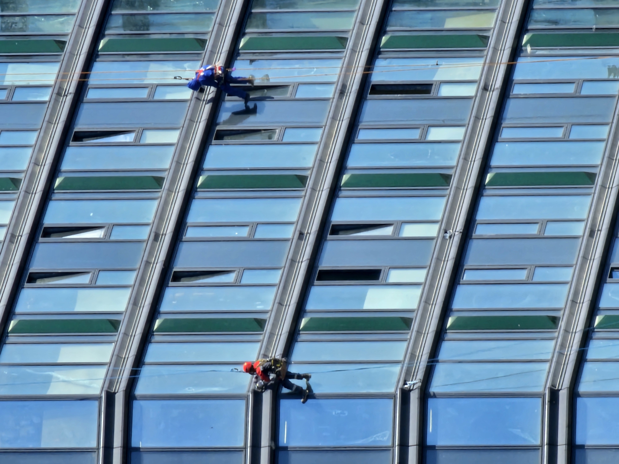 Two window cleaners are rappelling down the side of a modern glass-fronted building. One cleaner, wearing a red helmet and yellow and black gear, is on the left side of the frame, suspended by ropes. The other cleaner, dressed in blue, is higher up on the right side of the frame, also attached to ropes. The building has a grid-like pattern of blue and green tinted windows. The scene appears to be in an urban environment, possibly a business district, with the sun reflecting off the building's facade. There is no visual information to confirm the location as Moscow, Russia.