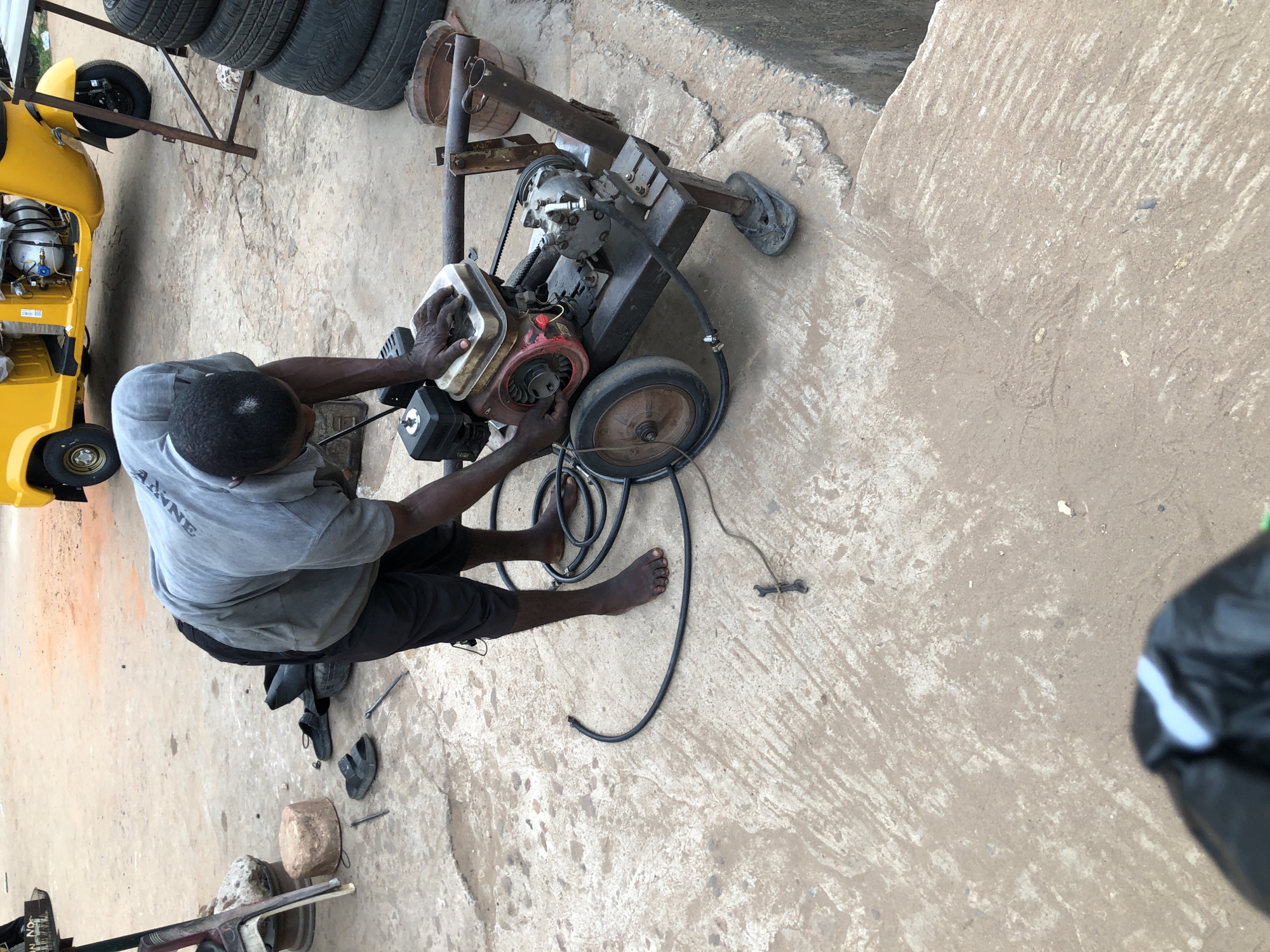 The image provides an overhead view of a man engaged in outdoor work, likely at a repair shop in Onitsha, Nigeria. The central subject is a man, seen from behind, wearing a grey t-shirt with the word "ANNE" visible on the back, and dark shorts. He is barefoot and appears to be concentrating on the task before him.
He is actively working on a small engine-powered machine, which includes a red and silver engine block, a small wheel, and several hoses and belts, possibly indicating it's a pressure washer, generator, or pump. His hands are positioned on different parts of the engine, suggesting he is performing maintenance or repair.
To the left of the man, a bright yellow tricycle (keke napep), a common mode of transport in Nigeria, is parked. A gas cylinder is visible within its open rear compartment. Further back and to the left, a stack of black tires is visible, along with a metal frame or stand. The ground is a rough, discolored concrete or cement surface, showing wear. Scattered around the man's feet are various tools and a pair of dark sandals. The scene is illuminated by natural daylight. A black plastic bag is partially visible on the right edge of the frame.