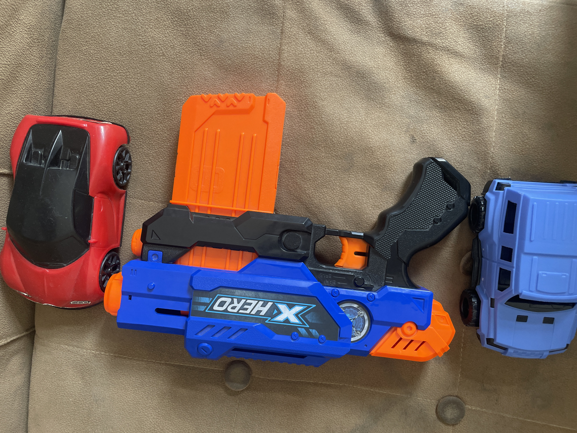 A red toy sports car is positioned on a textured brown surface. Below it, a blue and orange toy dart gun is centrally located. The dart gun has "X HERO" printed in white and blue on its side. Underneath the dart gun, a blue toy SUV is visible. The brown surface appears to be a couch or upholstered furniture. The image is taken from a top-down perspective. There is no visible indication of Tashkent, Uzbekistan or any specific urban location.