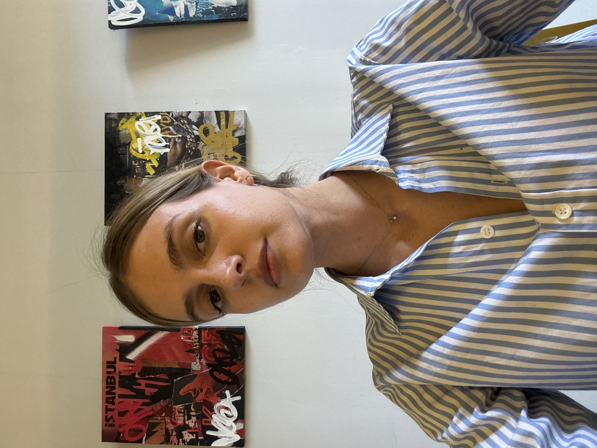 A young woman stands in front of a white wall adorned with three pieces of art. She wears a blue and white striped button-down shirt and a delicate necklace. The artwork consists of three small square canvases. One canvas features the word "ISTANBUL" in bold, red letters alongside other graffiti-style elements. Another canvas has a black background with splashes of yellow and white paint. The third canvas has a blue background with white abstract strokes. The scene takes place indoors in an unknown location in Tbilisi, Georgia.