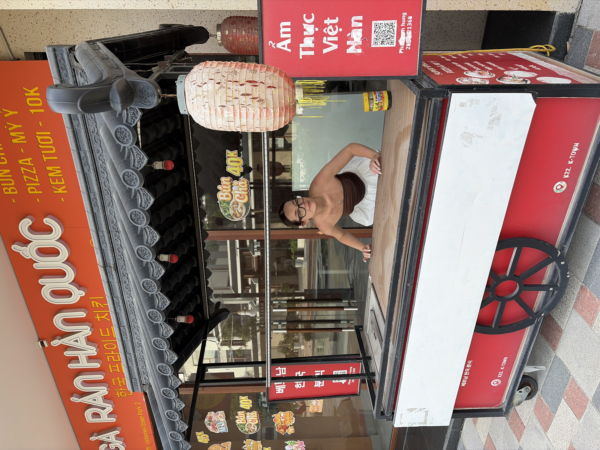 A young woman with glasses and dark hair, wearing a brown tube top and white skirt, stands behind a food cart under an overhang with a Korean-inspired roof. She is leaning forward, with her hands on the cart's counter. The food cart is red and white, with black accents and wheels. On the cart, there are various signs with Vietnamese and Korean text. One sign on the cart's side reads "K22. K-TOWN". The overhead sign displays "GA RÁN HÀN QUỐC" and Korean text. The background shows a street with buildings and trees. The scene is outdoors, likely in a commercial area.