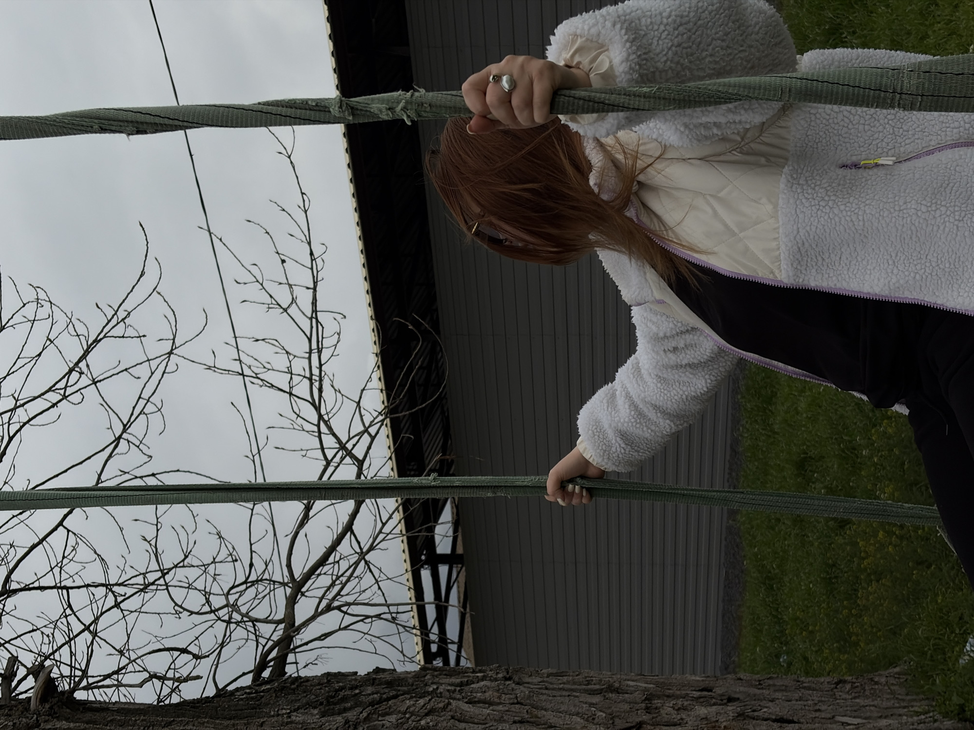 A young woman with auburn hair is hanging from a green rope or cable suspended between two trees. She is wearing a white, fuzzy jacket over a light-colored quilted vest, and a black shirt. The bottom of the frame shows the rough bark of a tree trunk, likely the base of the tree she's climbing. Behind her is a structure with corrugated metal siding. Bare branches are visible against an overcast sky. To the right, beyond the structure, is a patch of green vegetation. The image appears to be taken in a rural location. The location in the city of Port-Katon, Russia cannot be determined from the image.