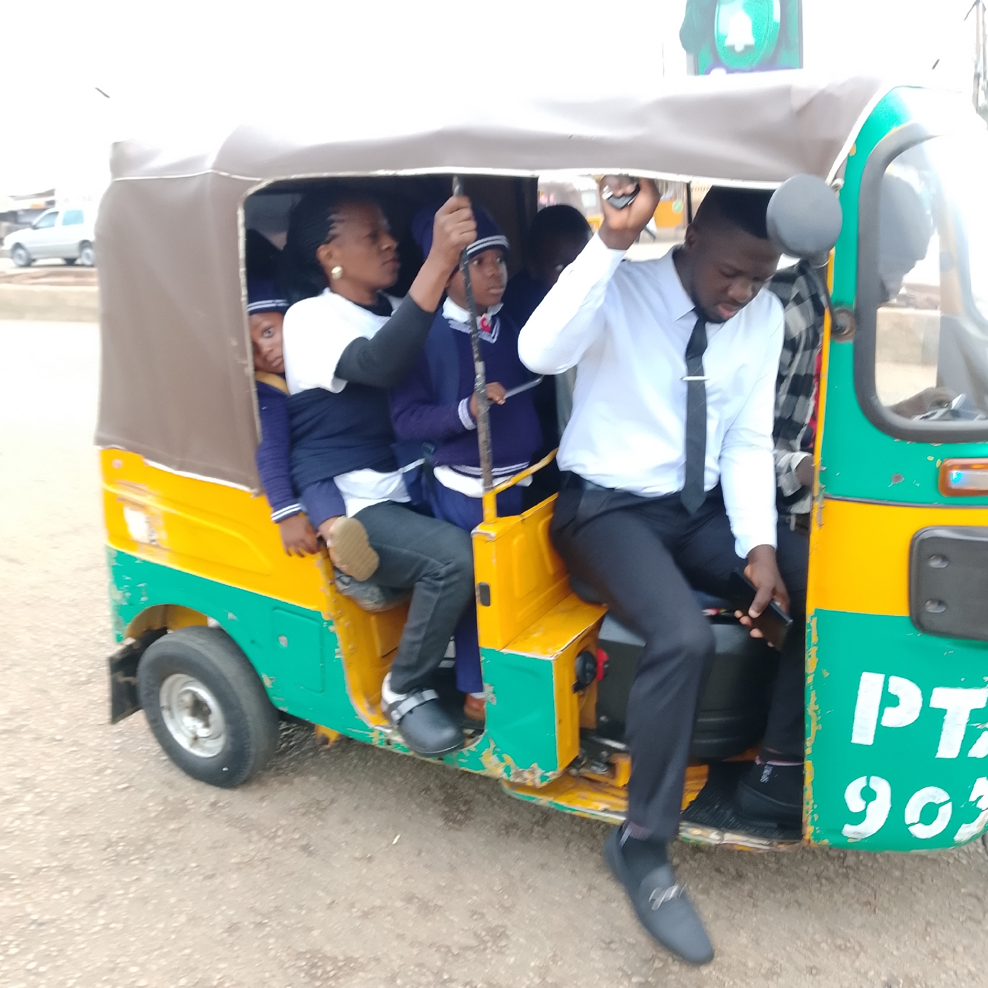 In an outdoor, unpaved urban environment, a group of people are packed into a three-wheeled auto rickshaw. The rickshaw is painted in bright green and yellow with some visible rust and wear. A man in a white collared shirt and black tie sits in the driver's seat, holding a mobile phone. Behind him, a person in a black and white plaid shirt is also visible. In the passenger area, a woman with braided hair and a white shirt is seated, with a young person in a blue school uniform next to her, gripping a metal bar. Another young person in a similar blue uniform is visible further back, peeking out. A white SUV is parked in the background. The image captures a moment of transit, with people of various ages sharing the confined space of the vehicle.