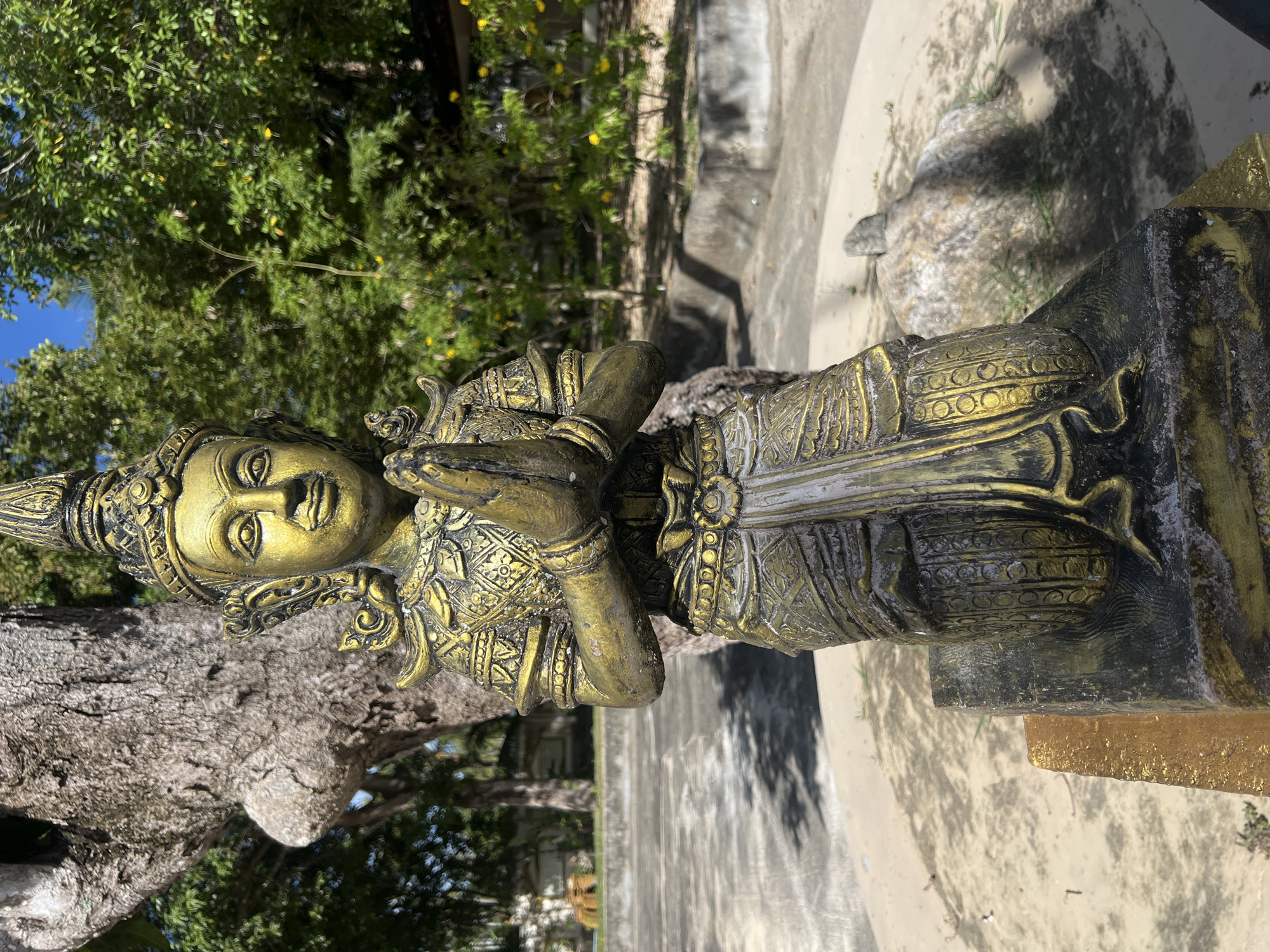 A golden statue of a person with hands clasped in a prayer-like gesture stands outdoors. The statue has ornate detailing on its clothing and headdress, with eyes wide open and a serene expression. It is positioned in front of a large tree with rough bark, and in the background, there are green trees with yellow flowers, a blue sky, and concrete structures. The lighting suggests it is daytime, likely sunny, with distinct shadows cast by the statue and surrounding elements. The statue appears to be a decorative element in a garden or temple setting, possibly in Ko Pha Ngan, Thailand, given the location context. There is no visible text in the image.