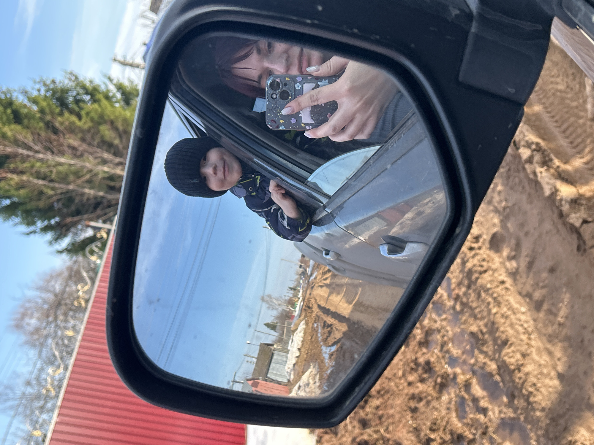 The image depicts a side-view mirror reflecting a scene with a child leaning out of a car window and a person holding a smartphone to take a photo. The reflection reveals the child in a black beanie and winter jacket, extending their arm out of the open window. Behind the child, a woman is visible, holding a smartphone with a floral case. Outside the car, the reflection shows a partly snowy landscape with trees and buildings. The exterior environment suggests a rural or suburban area, with a red metal fence visible in the background. The presence of snow indicates a cold season. The location is Фоки, Russia.