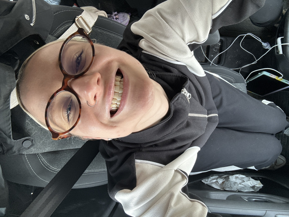 An adult individual with fair skin and light-colored hair, wearing tortoise-shell patterned glasses, a silver-colored hoop nose ring, and small stud earrings, is captured in a close-up, high-angle selfie. The individual is smiling broadly, revealing slightly yellowed teeth and an orthodontic retainer on the lower teeth, with crinkling around the eyes. The individual wears a black hooded sweatshirt with a silver-colored zipper and light beige/off-white panels on the shoulders and arms, black track pants with white side stripes, and white sneakers. The setting is inside a vehicle, likely a car, where the individual is seated in the front passenger seat. A black seatbelt is properly fastened across the chest. The dark gray upholstered car seat has white stitching. The car's floor is dark, with visible dust or debris, crumpled white paper, and tangled white electronic cables near an electronic device with a colorful case. The specific city location of Torbalı, Turkey, is not identifiable from the visual information provided within the image.