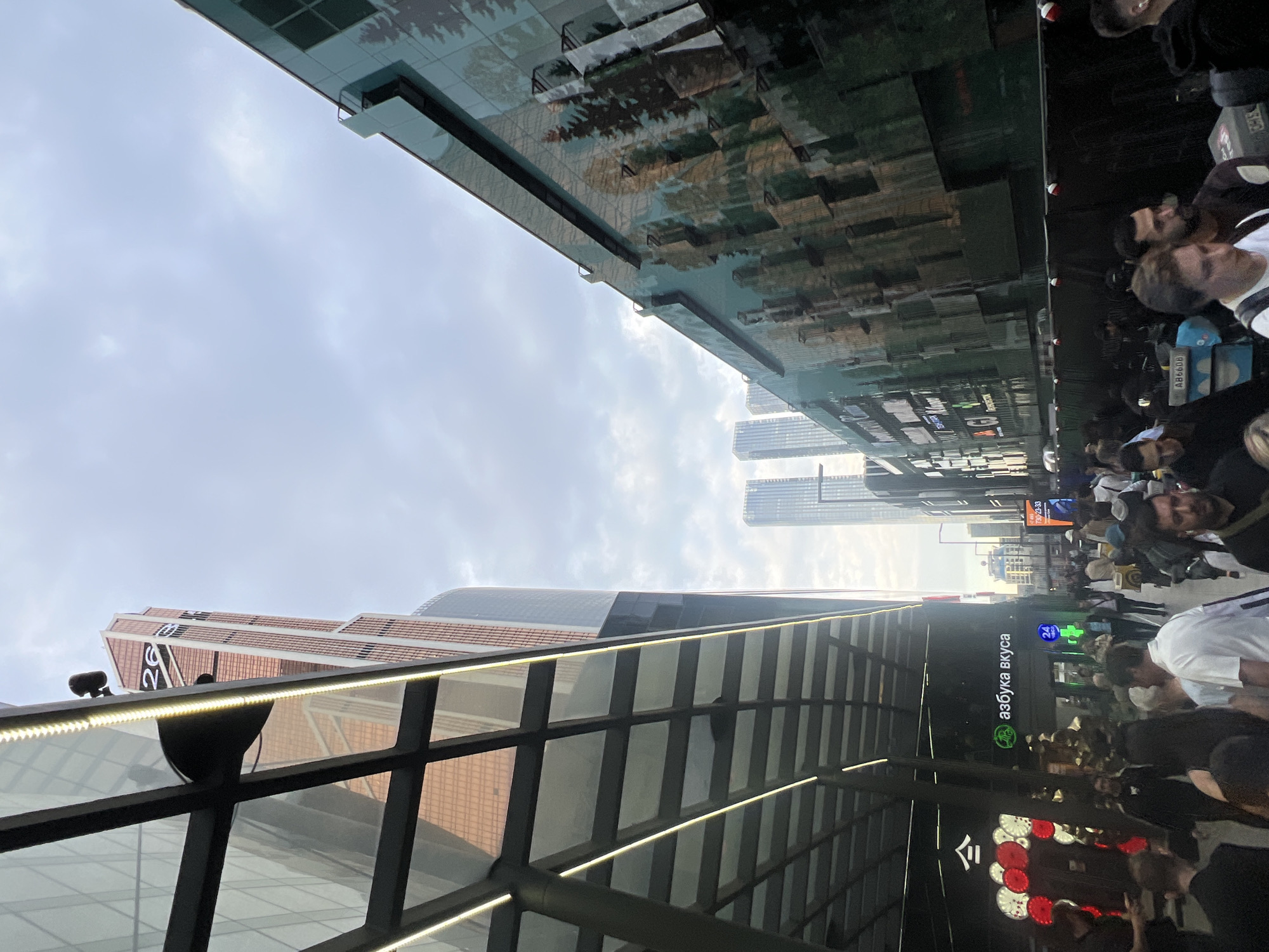 The image is a street-level perspective in a city with modern architecture, likely Khimki, Russia, based on the presence of a store sign reading "Азбука вкуса." Tall buildings are visible on either side of the street, reflecting in the glass surfaces. A pedestrian walkway or tunnel structure with a grid-like frame is visible in the foreground. A crowd of people walks along the sidewalk; their activities are not discernible. The sky is cloudy and overcast.