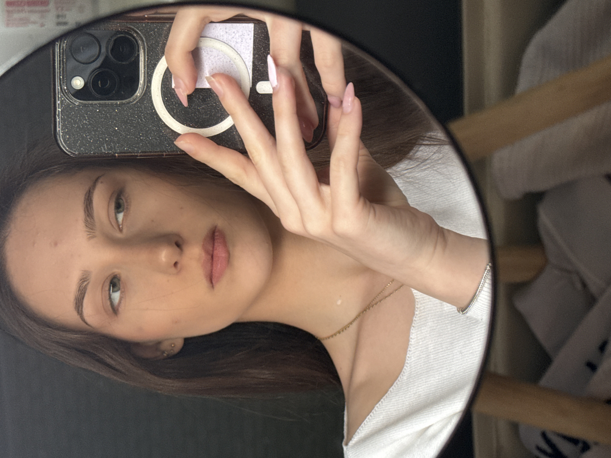 A woman is taking a selfie in a round mirror. She is holding a smartphone with a glittery case and a ring light attached. She has light skin, brown hair, and green eyes. She is wearing a white top and a gold necklace. The mirror reflects the background, which includes a black wall, a wooden chair, and some white clothing. The location cannot be determined from the image.