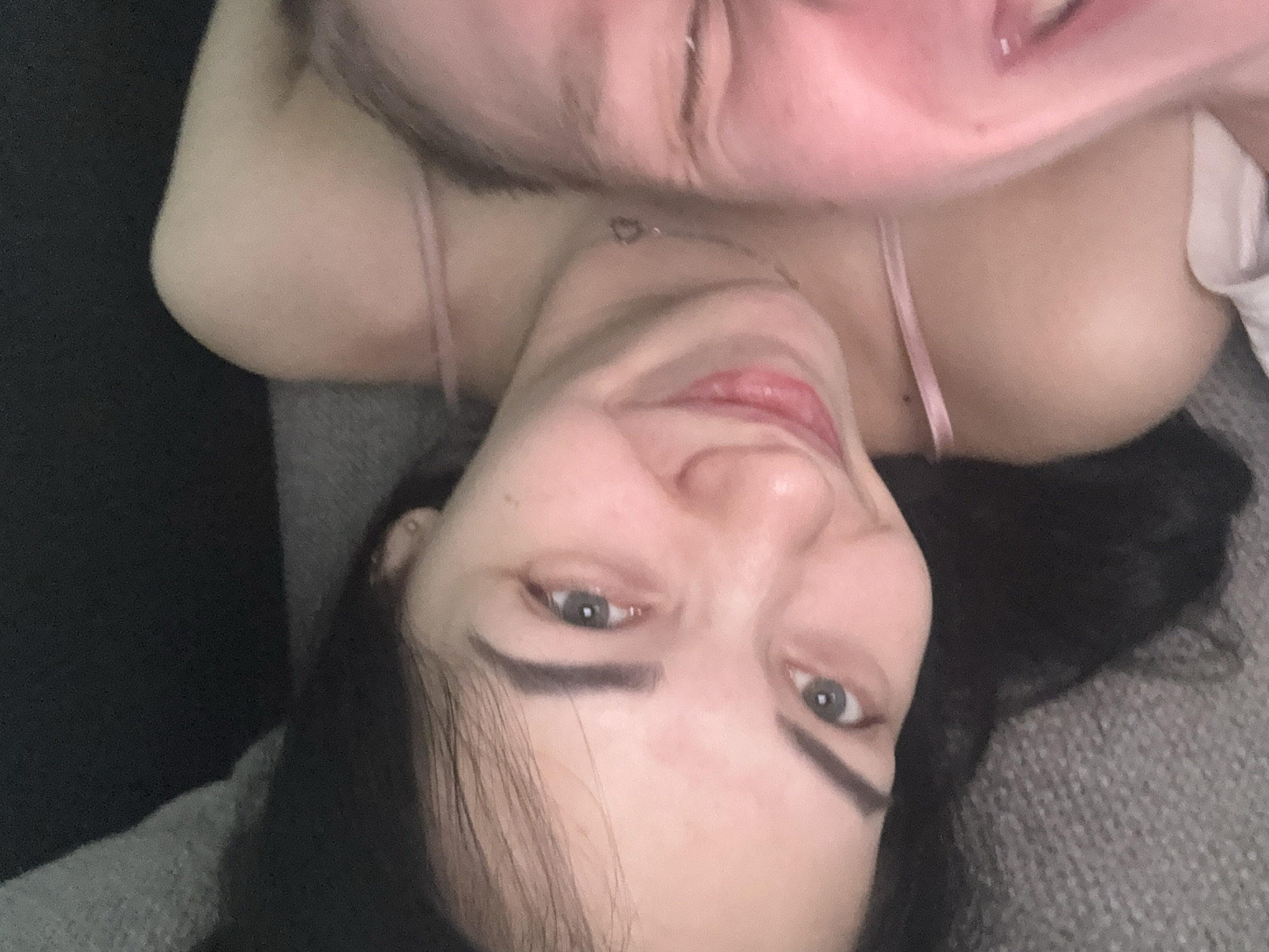 This is a close-up selfie of two individuals lying down, with one person's face prominently in the foreground. The person in the foreground has dark hair, light skin, and is looking towards the camera with a slight smile. Their eyes are open and appear to be a light color. They are wearing a pink strap that is visible on their shoulder. Another person is partially visible in the background, with their face out of focus and to the right. The background appears to be a couch or similar soft surface. The lighting is dim, suggesting an indoor setting.