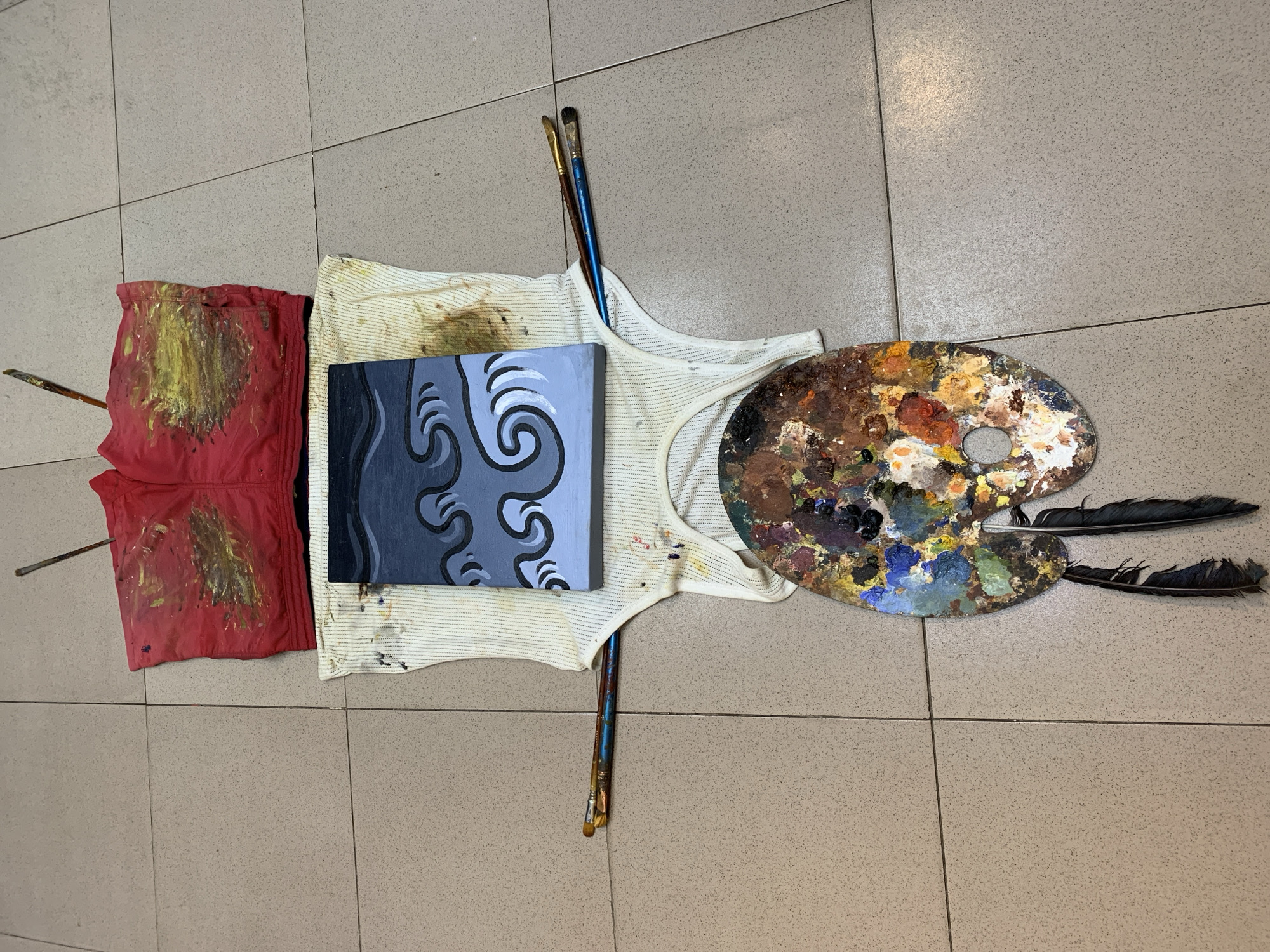 An overhead shot shows an art setup on a tiled floor, likely in Ado, Nigeria. A red garment, splattered with yellow paint, is at the top. Below it lies a white ribbed tank top, also stained with paint. Centered on the tank top is a small canvas painting of dark blue and gray waves. Two paintbrushes with blue handles are placed horizontally across the tank top. Beneath the tank top is a wooden artist's palette, covered in a wide array of colorful paints, with two dark feathers hanging from its lower edge. The scene appears to be an artist's workspace, possibly a studio or an outdoor area.