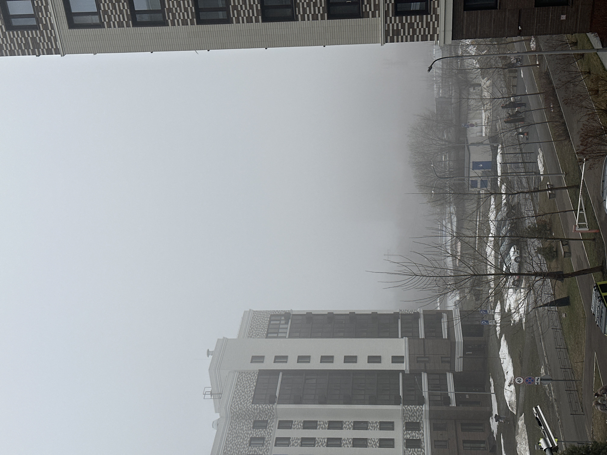 The image depicts a foggy cityscape, likely in Kazan, Russia, showing buildings, trees, roads, and patches of melting snow. A tall building with a patterned facade is partially visible on the left side. Another building of similar design, with rows of windows, appears at the bottom. In the middle ground, bare trees line a road where some workers are working and melting snow patches persist. Streetlights are spaced along the road. The heavy fog obscures the background, creating a hazy and muted atmosphere. A parking sign and speed limit sign are visible near the lower-right corner of the image.