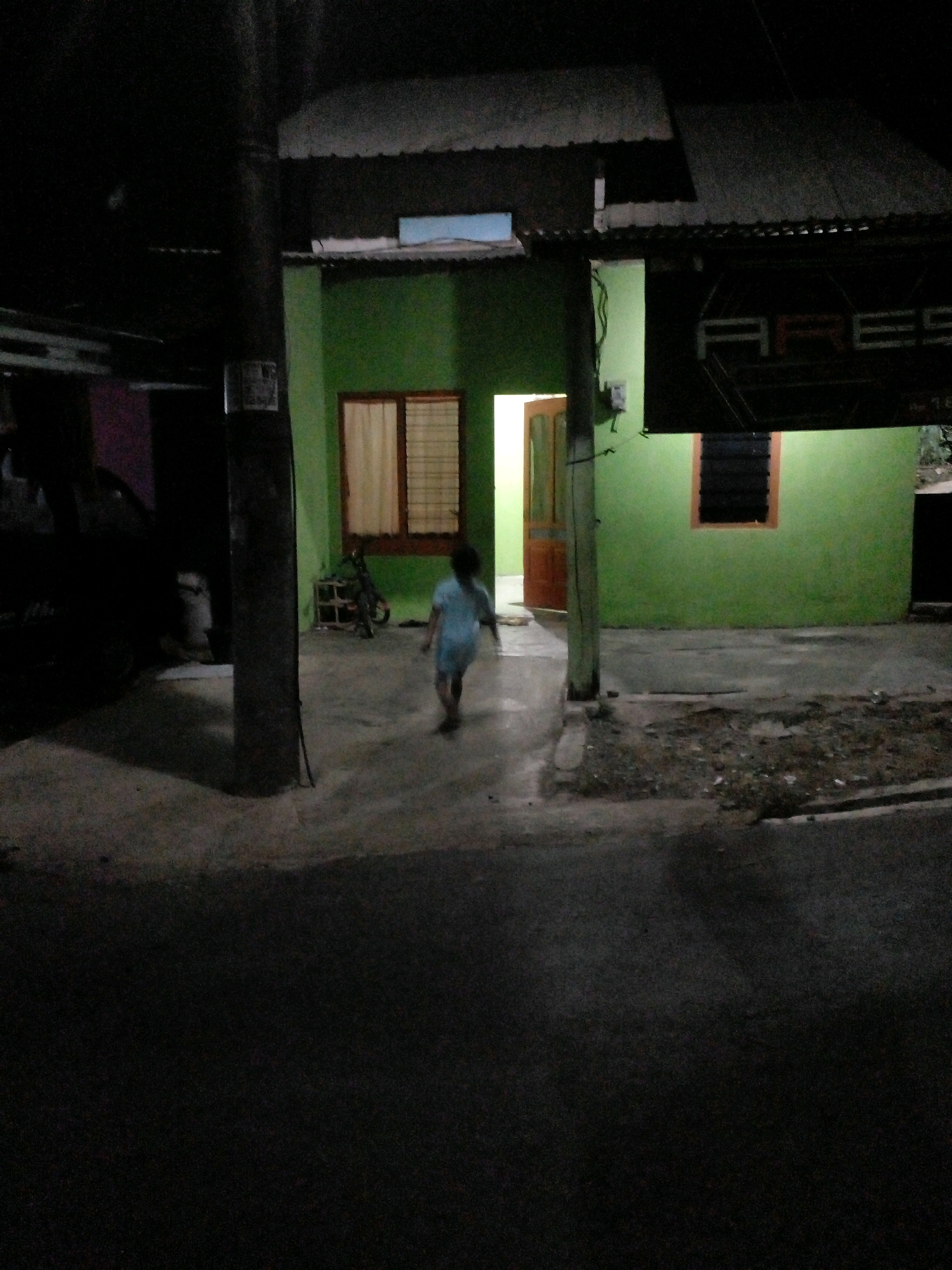 A low-light, slightly blurry, full-frame shot captures a young child in a light-colored dress running away from the camera on a paved area in front of a green building. The building has a dark-colored roof and a wooden-framed window with a light-colored curtain to the left, and a dark-slatted window to the right. An open wooden door reveals a bright interior. A utility pole with electrical wires stands to the left of the frame. In the background, a bicycle is visible. The ground in front of the building is a mixture of dirt and pavement, with debris scattered on the surface. The foreground is dark pavement. The scene appears to be a residential area in Pungangan, Indonesia.