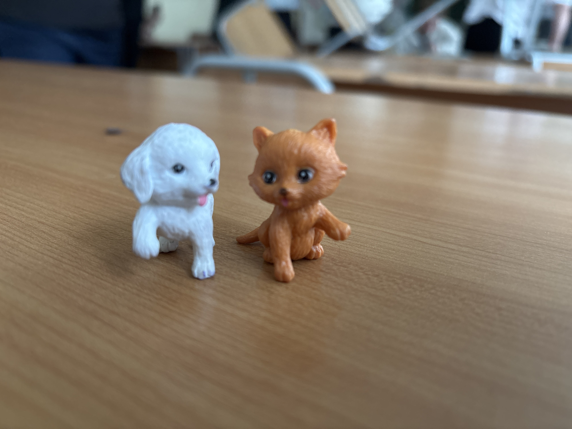 The image displays two small plastic figurines, a white dog and an orange cat, positioned side-by-side on a light brown wooden surface, likely a desk or table. The white dog figurine, on the left, has textured fur, dark eyes, and a small pink tongue sticking out. Its front paws are slightly raised, and its back paws show subtle purple markings. The orange cat figurine, on the right, is a solid color with defined ears, large dark eyes, and a small nose, looking directly forward. It is in a sitting or crouched pose with its front paws outstretched.
The background is significantly out of focus, rendering it blurry and indistinct. It appears to be an indoor setting with muted colors and vague shapes that suggest furniture, possibly chairs or other tables, indicative of a room environment like a classroom or office. The wooden surface has a subtle grain pattern. No text is visible in the image. The lighting is soft and even, suggesting indoor illumination without harsh shadows.