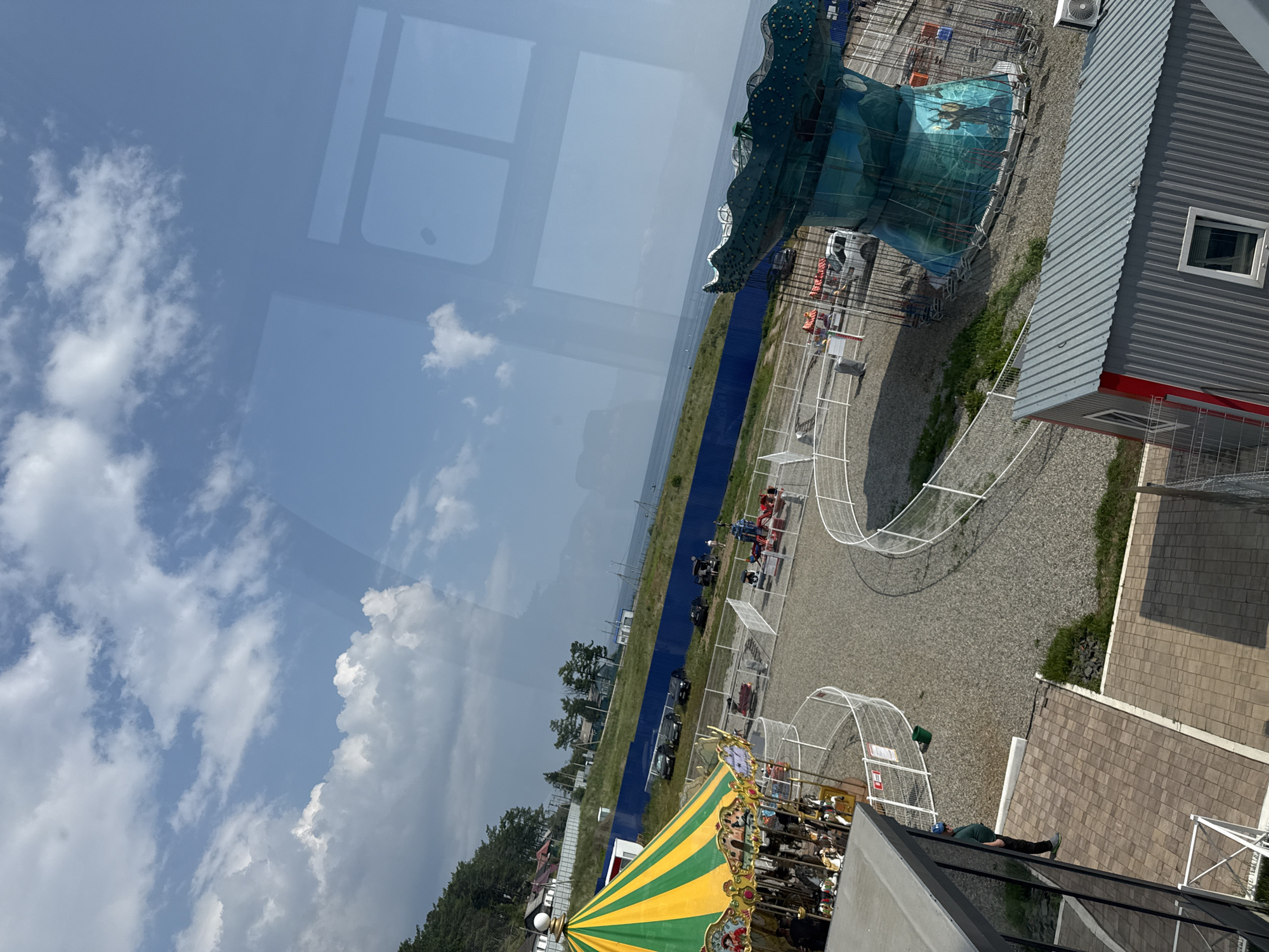This is a high-angle, wide shot of an amusement park in Listvyanka, Russia, on a clear day with scattered clouds. In the foreground, a person in a green shirt and dark pants stands on a tiled patio, looking to their left. To the right of the person, a corrugated metal building with red trim and a small window is visible.
Further into the park, a yellow and green striped carousel is partially visible on the left. A blue fence runs horizontally across the middle ground, separating the park from a grassy area and a body of water, likely Lake Baikal. Beyond the fence, several cars are parked.
In the center of the park, a ride with a large, teal, wave-shaped structure with a mural depicting a Viking is prominent. Several smaller amusement rides are scattered throughout the park, enclosed by white chain-link fences. The ground of the park is covered in gravel. The sky is blue with fluffy white clouds. The overall impression is a sunny day at a small amusement park with a view of the water.