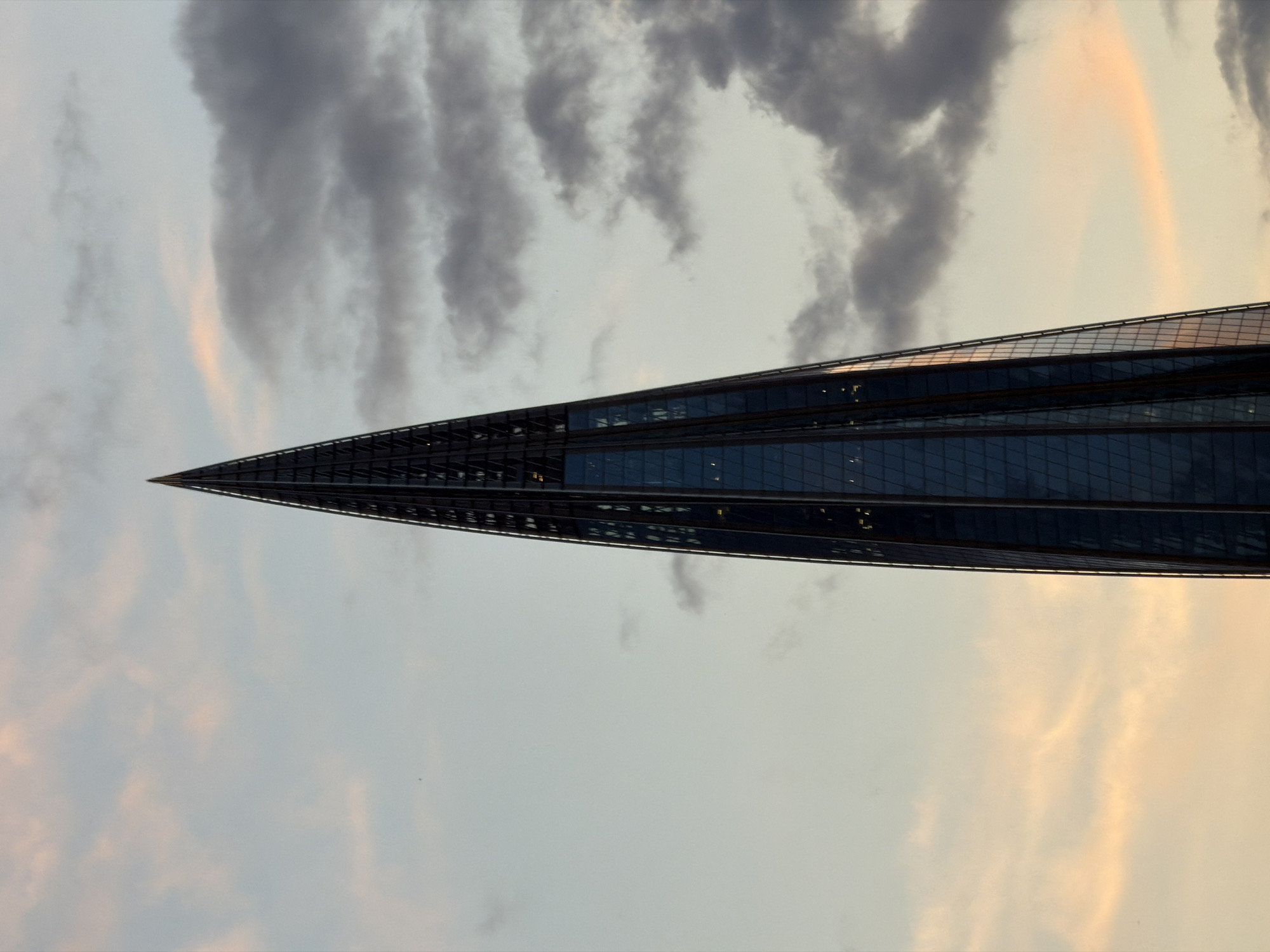The image features a striking, modern skyscraper, likely the Lakhta Center, dominating the frame with its slender, tapering form and reflective glass facade. The building extends diagonally from the lower right to the upper left, showcasing its sharp, pointed peak. The sky behind the structure is a prominent element, filled with a dynamic mix of clouds, ranging from dark, heavy grey formations to lighter, more diffused patches. Soft, warm hues of pale orange and pink are visible in parts of the sky, particularly towards the upper right and lower left, suggesting either sunrise or sunset. A faint but discernible rainbow arc is present in the upper right portion of the sky, positioned behind the building. There is no visible text or specific activity taking place. The atmospheric conditions, including the varied cloud cover and rainbow, imply recent rain or atmospheric moisture. The setting is Saint Petersburg, Russia.