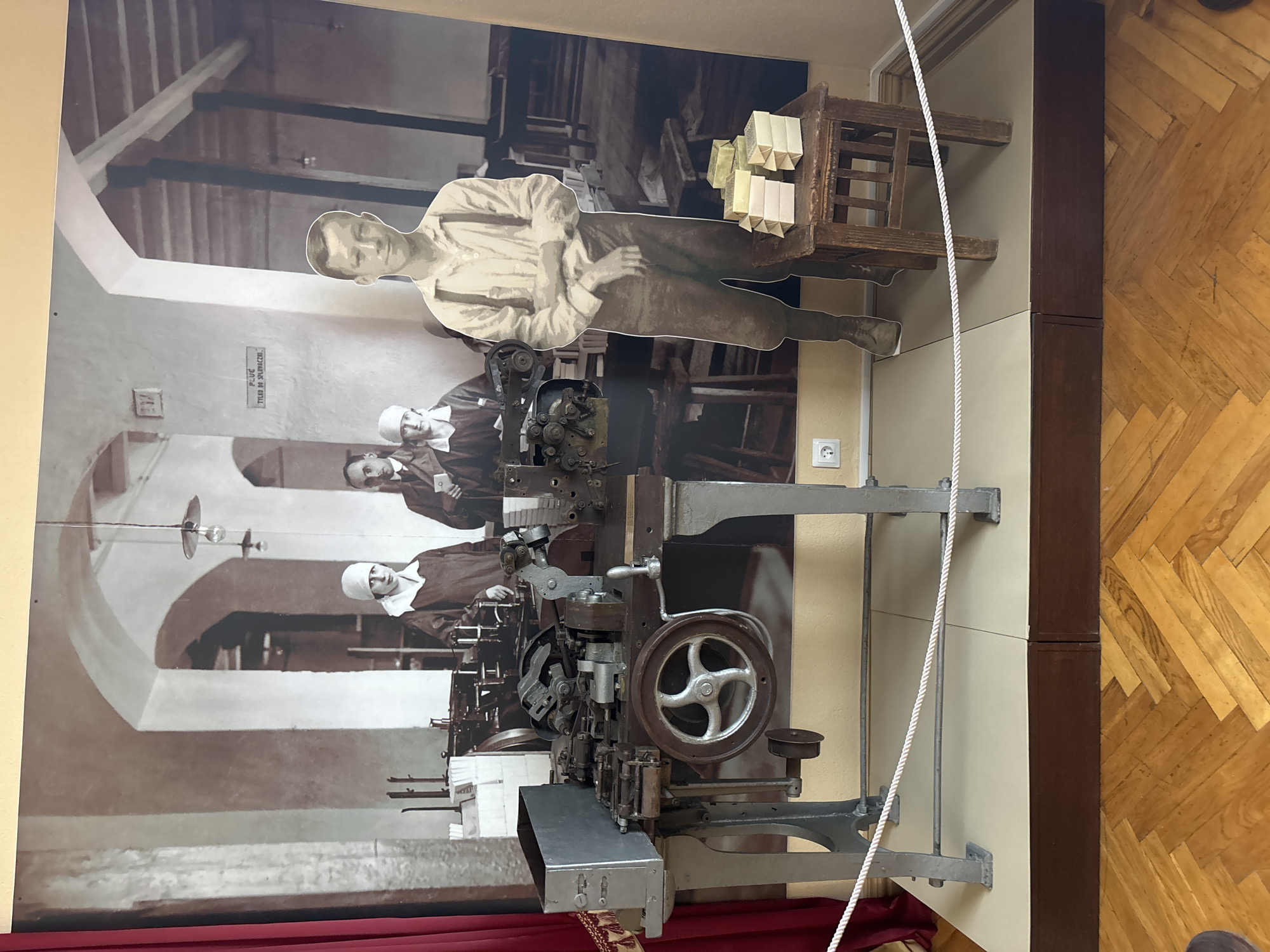 The image depicts a display in a museum in Hrodna, Belarus, likely related to industrial history or a specific factory. The central focus is a large photographic mural of a historical factory setting, showing workers in sepia tones. In front of this mural, a life-sized cardboard cutout of a young boy wearing suspenders stands to the right, with a small wooden stool beside him. On the stool are several stacks of light-colored rectangular objects, possibly soap or packaged goods. To the left of the mural, a vintage industrial machine, likely a manufacturing device with gears, wheels, and metal components, is prominently displayed on a grey metal stand. The machine has a large, spoked wheel on its front. A silver metal box is attached to the left side of the machine. A red curtain hangs on the far left of the scene. The display is set on a platform raised from the wooden parquet floor. A white rope barrier is in place in front of the display. The overall environment suggests an indoor exhibition space.