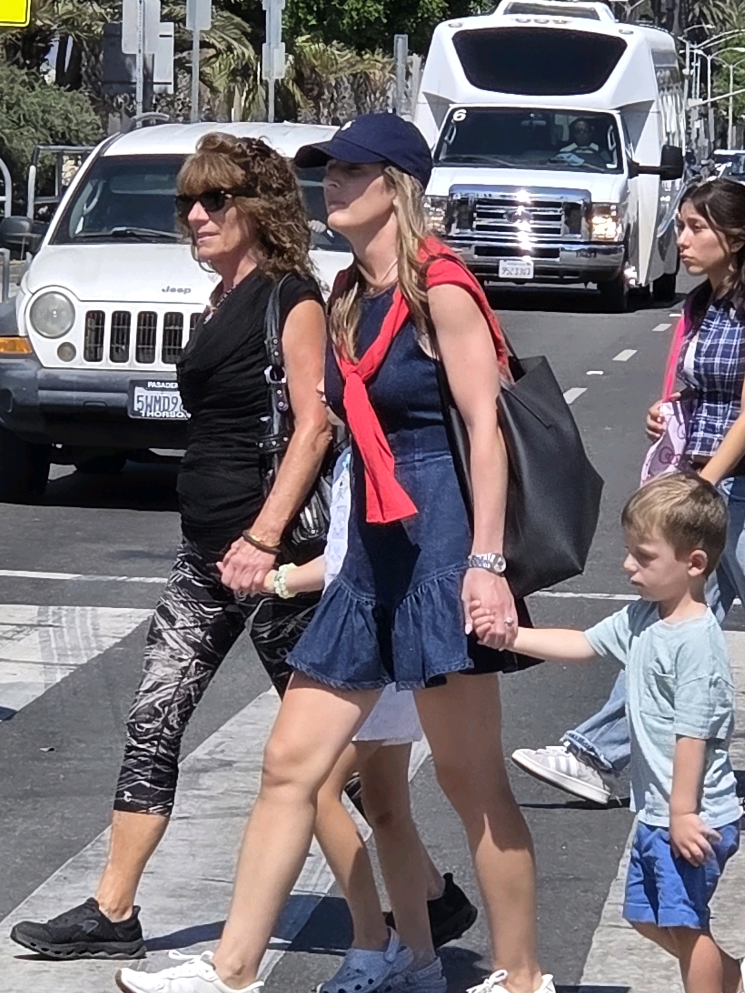 An outdoor, eye-level, medium shot captures a group of people crossing a street on a sunny day in Santa Monica, California, United States of America. In the foreground, a woman with curly brown hair and sunglasses, wearing a black sleeveless shirt and patterned leggings, holds hands with a young boy in a light blue t-shirt and blue shorts. The boy is also holding hands with another woman in a denim dress and a red scarf draped over her shoulders. Behind them, a younger woman in a blue and white plaid shirt and a white shirt underneath is visible, and further back, a white shuttle bus with tinted windows is driving down the street, with a driver in the front seat. A white Jeep is parked to the left of the shuttle bus. The ground appears to be a crosswalk with white stripes, and palm trees are visible in the background.