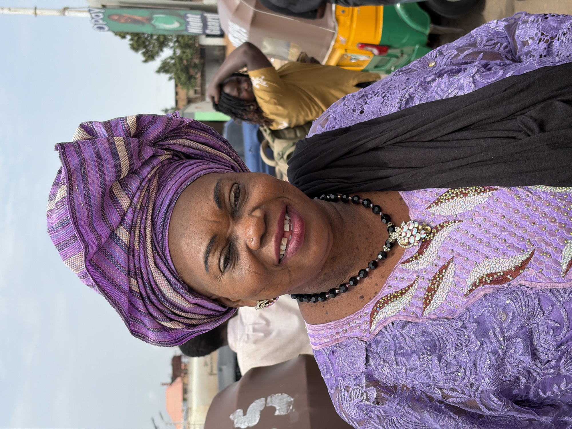 A woman in a purple and beige striped turban and a purple lace dress stands smiling in an outdoor urban environment. She wears a black shawl and a black beaded necklace with a jeweled pendant. Behind her, a man in a mustard-colored shirt touches his head while sitting in a yellow and green vehicle. A sign that reads "Night Guard" is visible in the upper left corner. The location appears to be the city of Gura Top, Nigeria.