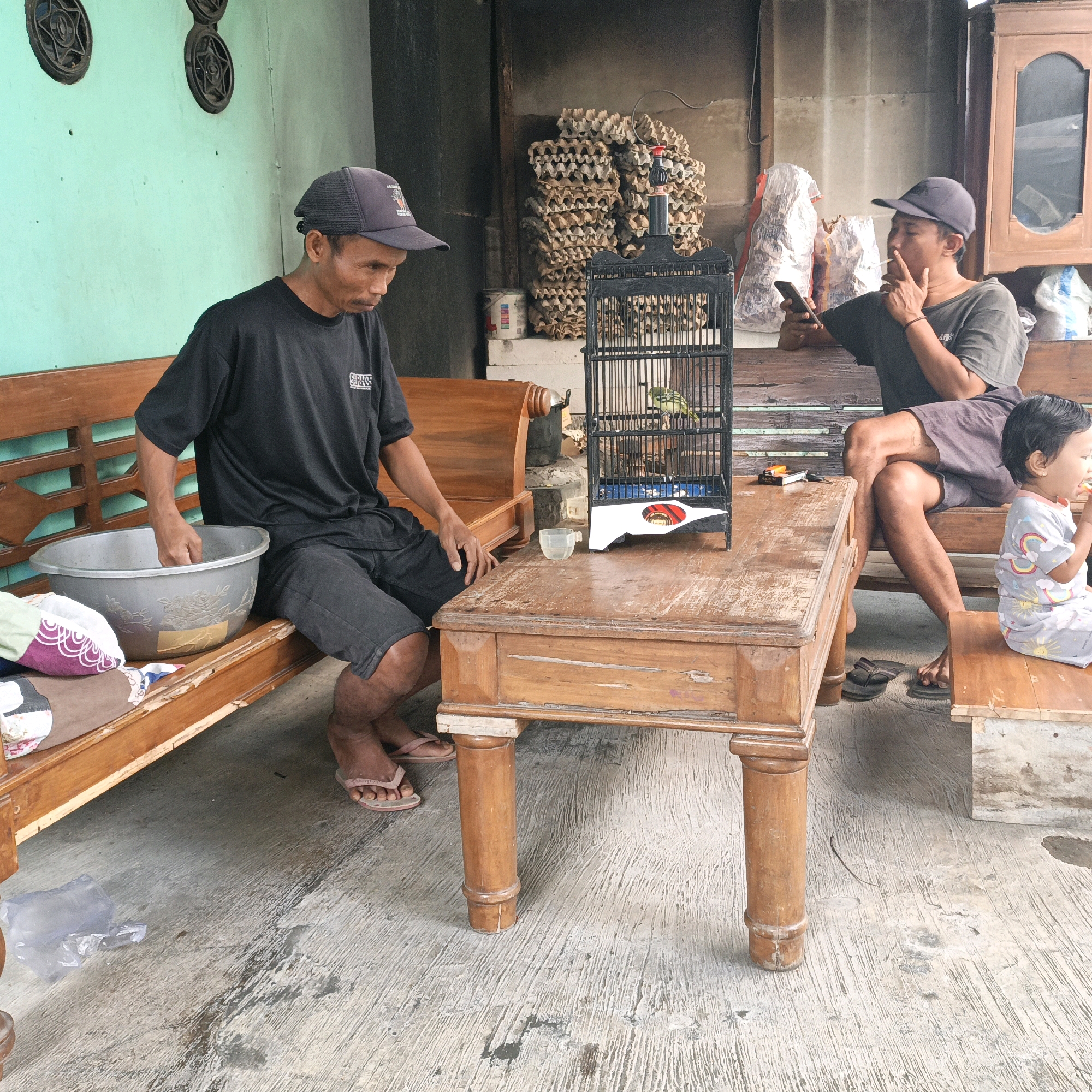 In Tanahbaya, Indonesia, a man wearing a dark baseball cap and a black t-shirt sits on a wooden bench. In front of him, on a wooden table, is a black birdcage containing a small green bird. To his right, another man in a dark shirt and baseball cap is smoking a cigarette and looking at his phone, also seated on a wooden bench. A young child in a grey shirt with colorful rainbow and sun motifs is seated to the right of the second man, facing away from the camera and eating. The environment appears to be an outdoor area with a concrete floor, a green wall with star-shaped decorative elements on the left, and stacks of what appear to be egg cartons in the background. A large metal basin is on the bench next to the first man.