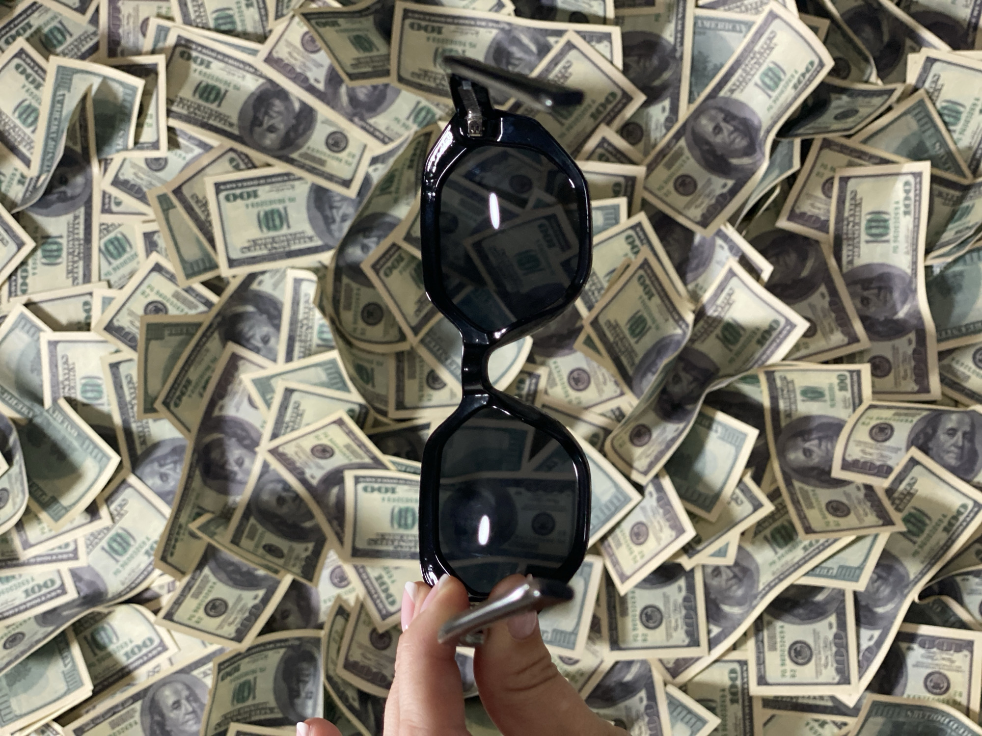 The image depicts a hand holding up a pair of black sunglasses. The lenses of the sunglasses reflect the surrounding scene. The background is filled with scattered US one hundred dollar bills. The image provides no information to determine a location in Sochi, Russia.