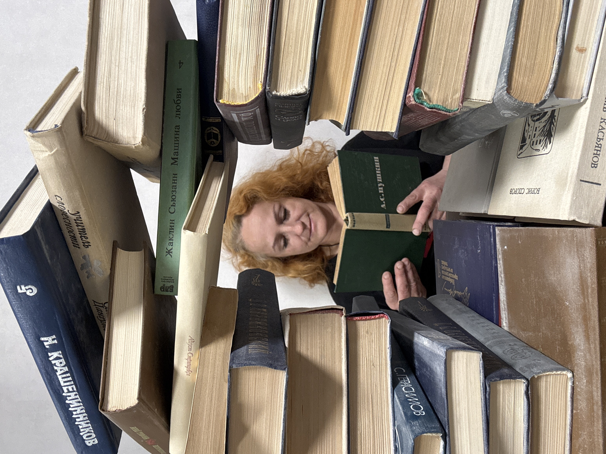 The image presents a woman engaged in reading, framed by a dense, almost architectural arrangement of stacked books. Her reddish-blonde hair and smiling face are visible through an opening created by the books, with her hands holding an open volume. The perspective is from above, looking down into this literary 'nest'.
The books, varying in color, size, and age, are piled in an eclectic manner, some upright, some lying flat, and others tilted, creating a dynamic and somewhat chaotic structure around the reader. Many books show signs of wear, with yellowed pages and faded spines. The background behind the woman within the book enclosure is a plain, light surface.
The woman's expression is one of calm concentration, suggesting deep immersion in her reading. Visible text on the books is predominantly in Cyrillic script. Notable titles and authors include "А.С.Пушкин" (A.S. Pushkin) on a dark green book, "Жаклин Сьюзанн Машина любви" (Jacqueline Susann Machine of Love), "КАСЬЯНОВ" (KASYANOV), "БОРИС СПОРОВ" (Boris Sporov), "С. ГЕРАСИМОВ" (S. Gerasimov), "Август Стриндберг" (August Strindberg), and "Н. КРАШЕНИННИКОВ" (N. KRASHENINNIKOV). Other phrases like "учитель словесности" (teacher of literature/philology) and "в четырех произведения" (in four works) are also discernible. The extensive Cyrillic text aligns with the provided location context of Chaikovsky, Russia.