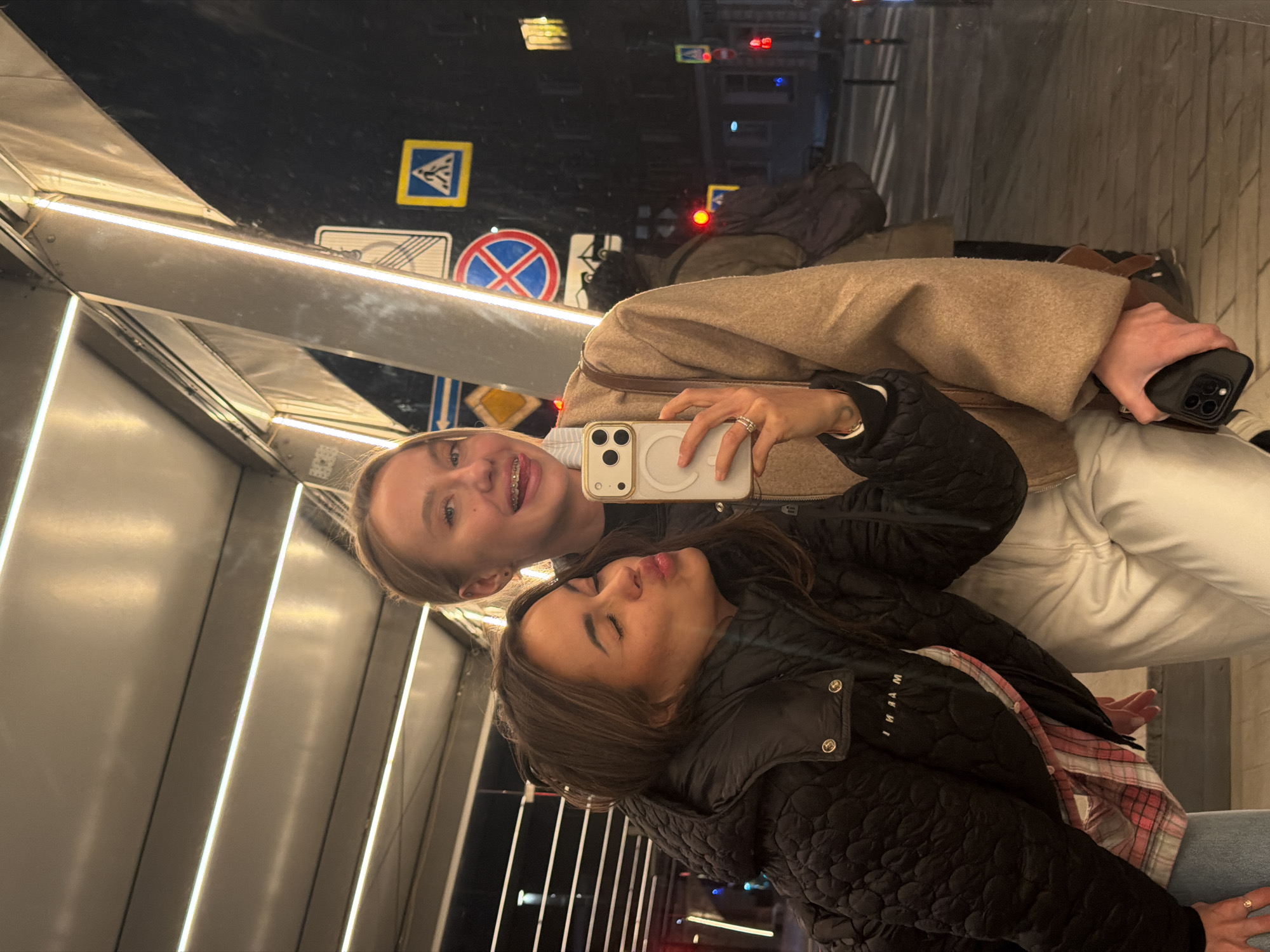 Two women are taking a selfie in a mirrored pedestrian overpass in Krasnoyarsk, Russia. The blonde woman on the upper left is holding a white iPhone and wearing braces. The brunette woman is kissing in the lower portion of the image. Behind the women, the mirror reflects street signs, traffic lights, and blurred figures. The overpass features a modern design with bright linear lighting.