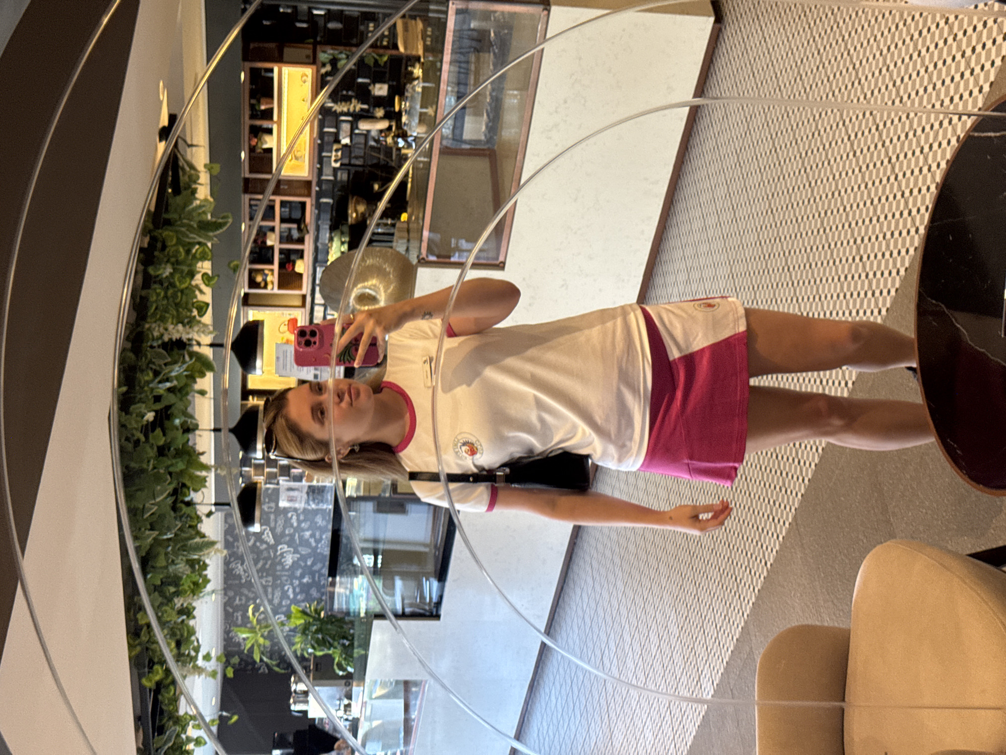 A young woman is standing in front of a large mirror, taking a selfie with her phone. She is wearing a white t-shirt with pink trim and a matching pink skirt. She has a black purse slung over her shoulder. The mirror reflects a cafe interior with plants, shelves, and display cases. The woman's reflection is the primary subject of the image. The location appears to be a modern cafe.