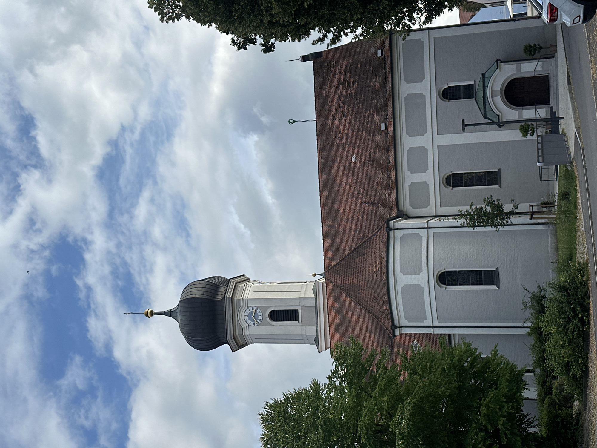 The image shows a church in Au in der Hallertau, Germany. The church has a tall, grey tower with a dark, onion-shaped dome topped with a cross. A clock face is visible on the tower. The main body of the church is also grey with a red-tiled roof. There are arched windows with stained glass and a main entrance with a green-domed canopy. A large tree with green foliage stands to the left of the church. The sky is partially cloudy with some blue sky visible. In the foreground, there is a sidewalk and a street. A white car is partially visible on the right side of the image. A small bird is flying in the sky.