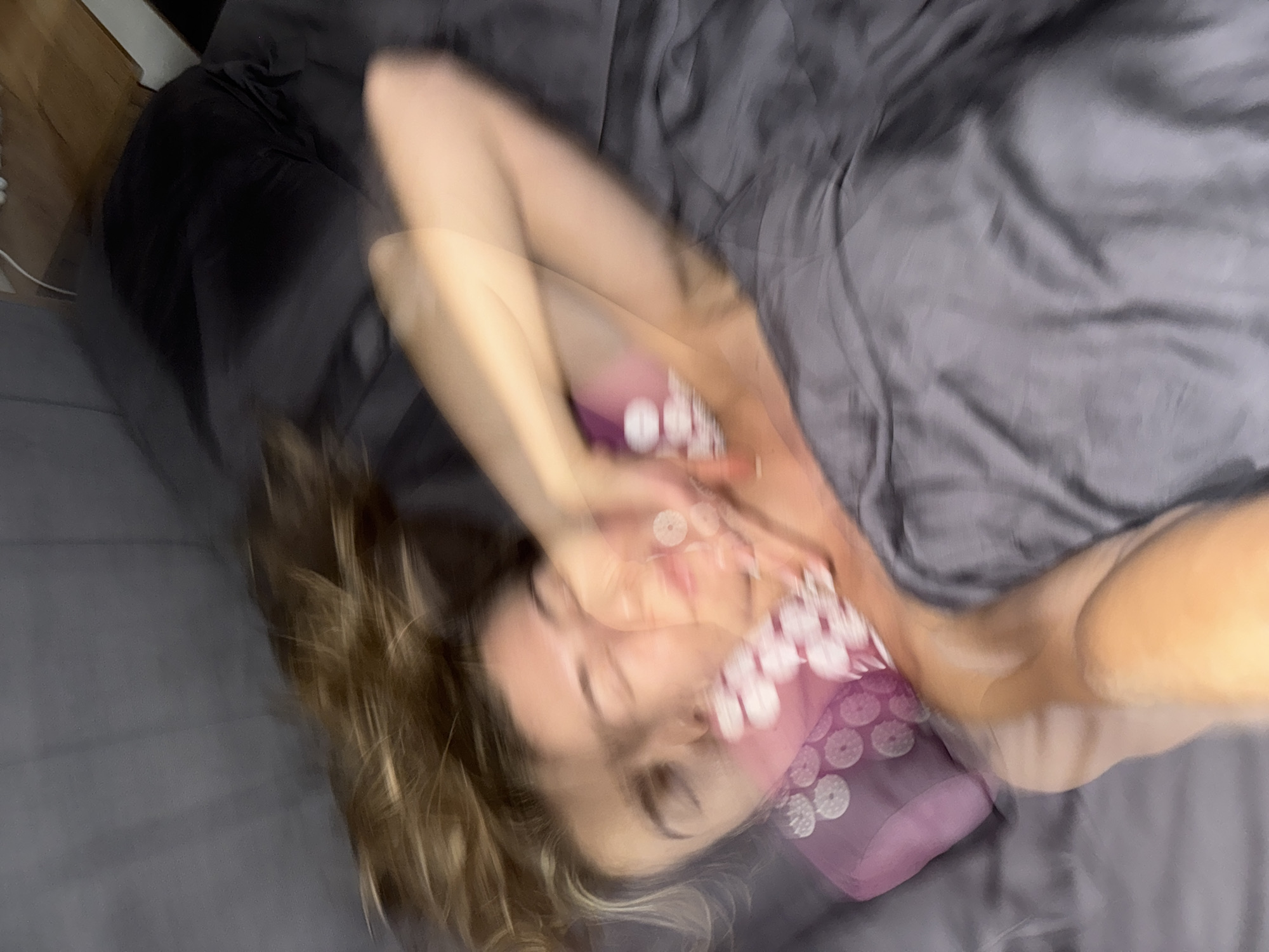 An overhead shot shows a person lying in a bed with gray sheets. The person has blonde hair and appears to be reaching up towards their face. A pink acupressure mat is visible under their head and neck. The image is blurred due to motion, creating a double-exposure effect.