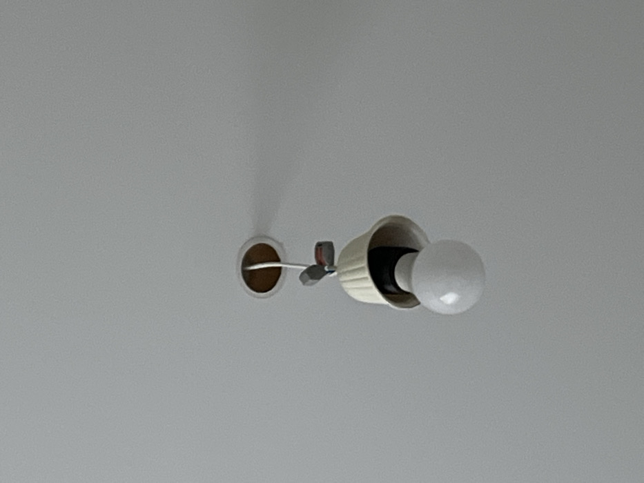 An indoor scene in Zainsk, Russia, depicts a partially dislodged or damaged ceiling light fixture against a plain, light-colored ceiling. A spherical white light bulb is screwed into a visible black socket. Above the socket, a cream-colored, bell-shaped light shade with vertical ridges hangs unevenly, exposing the internal components. White electrical wiring descends from a circular opening in the ceiling, revealing the brown edge of the aperture. Two small, grey electrical wire connectors are visible on the wiring segment above the shade, one featuring a red marking. No individuals are present in the frame.