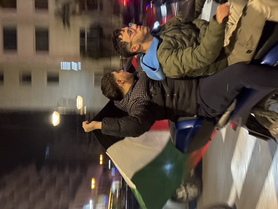 Two young men ride a blue motorcycle at night. The first man, in an olive green jacket, khaki pants, and a blue hoodie, looks up and to the right. The second man, wearing a black puffer jacket over a checkered shirt, holds a Palestinian flag in his right hand. Streetlights illuminate the background, revealing buildings along the road. The image has a slightly blurred effect, suggesting movement.