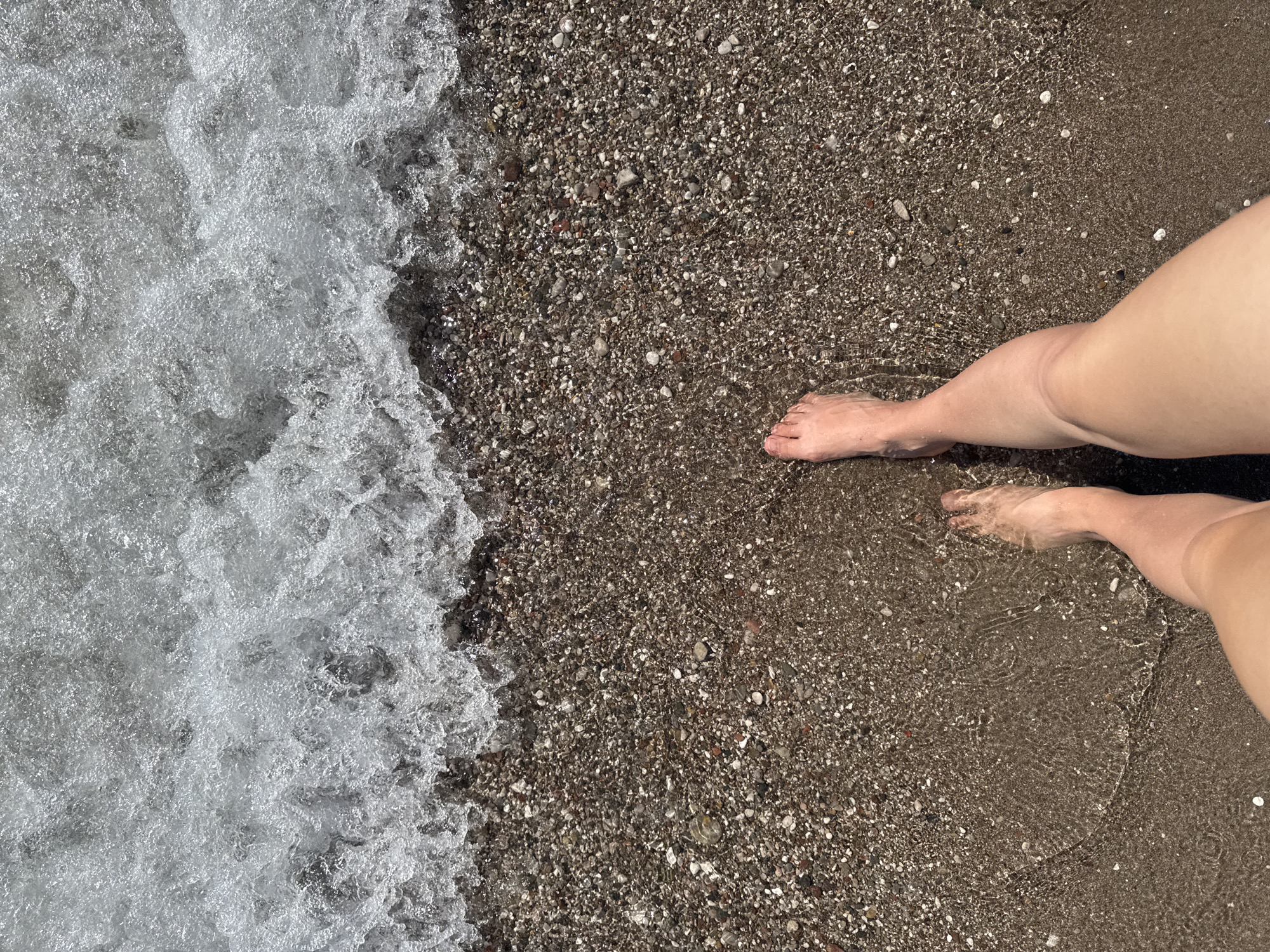 In Šušanj, Montenegro, a close-up shot captures the lower legs and feet of a person standing on a pebble beach. The water's edge is visible, with a small wave receding from the shore. The pebbles are varied in color and size, with some appearing wet and dark, while others are lighter and dry. The water is clear, allowing the pebbles beneath to be seen. The person's skin is fair, and their feet are bare. The scene is illuminated by natural daylight, creating a bright and airy atmosphere.