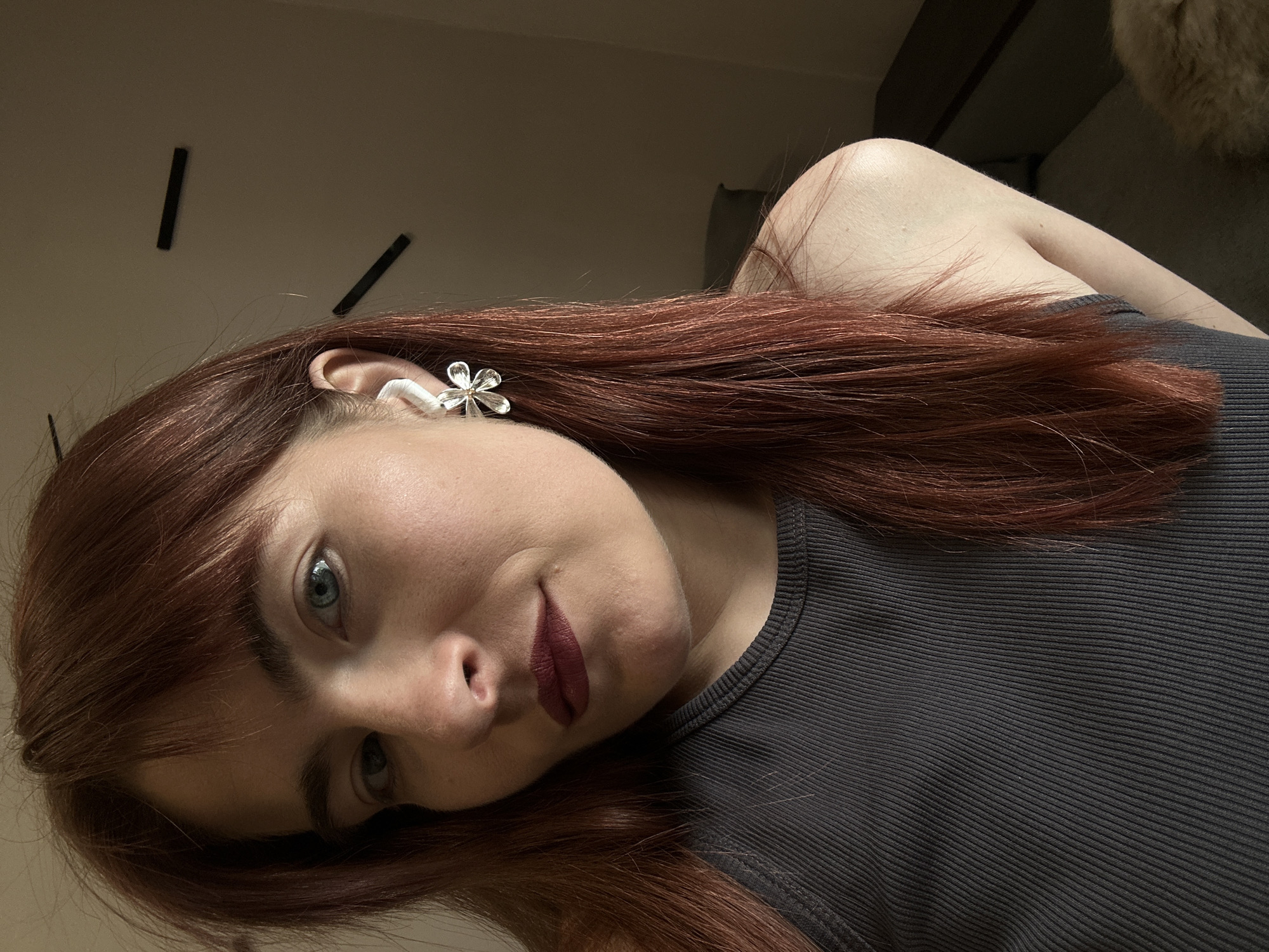 A woman with light skin and red hair is positioned in the foreground, tilted to the side. She wears a gray ribbed top and a silver flower earring. Her makeup includes dark lipstick. The background is out of focus, but reveals portions of a beige wall with black rectangular accents. The location is indeterminable from the image. The city of Kuda, Russia, cannot be confirmed from the information available in the image.