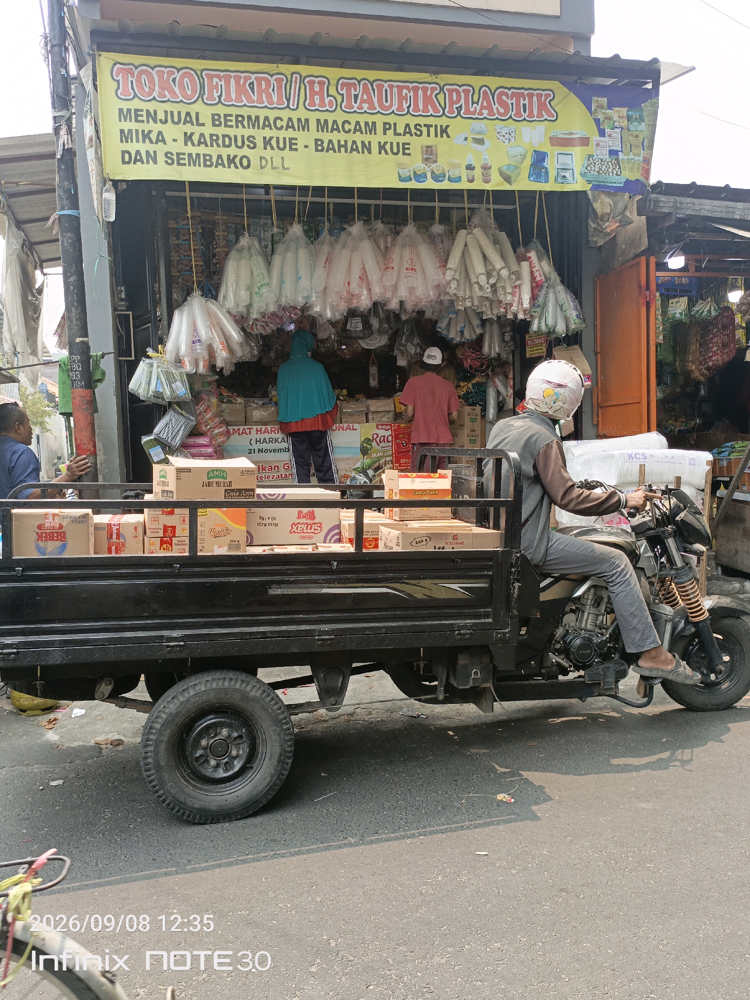 A man wearing a helmet with a cartoon character on it is riding a black three-wheeled motorcycle with a flatbed loaded with cardboard boxes. He is wearing a gray long-sleeved shirt and gray pants, and his right foot is on the footrest. The motorcycle is parked on a street in front of a shop with a yellow banner that reads "TOKO FIKRI / H. TAUFIK PLASTIK". The shop sells various plastic items, cake boxes, and groceries. Two women are standing inside the shop, one wearing a teal hijab and the other a pink shirt. The street is paved and appears to be in a commercial area. The image was taken on September 8, 2026, at 12:35 PM.