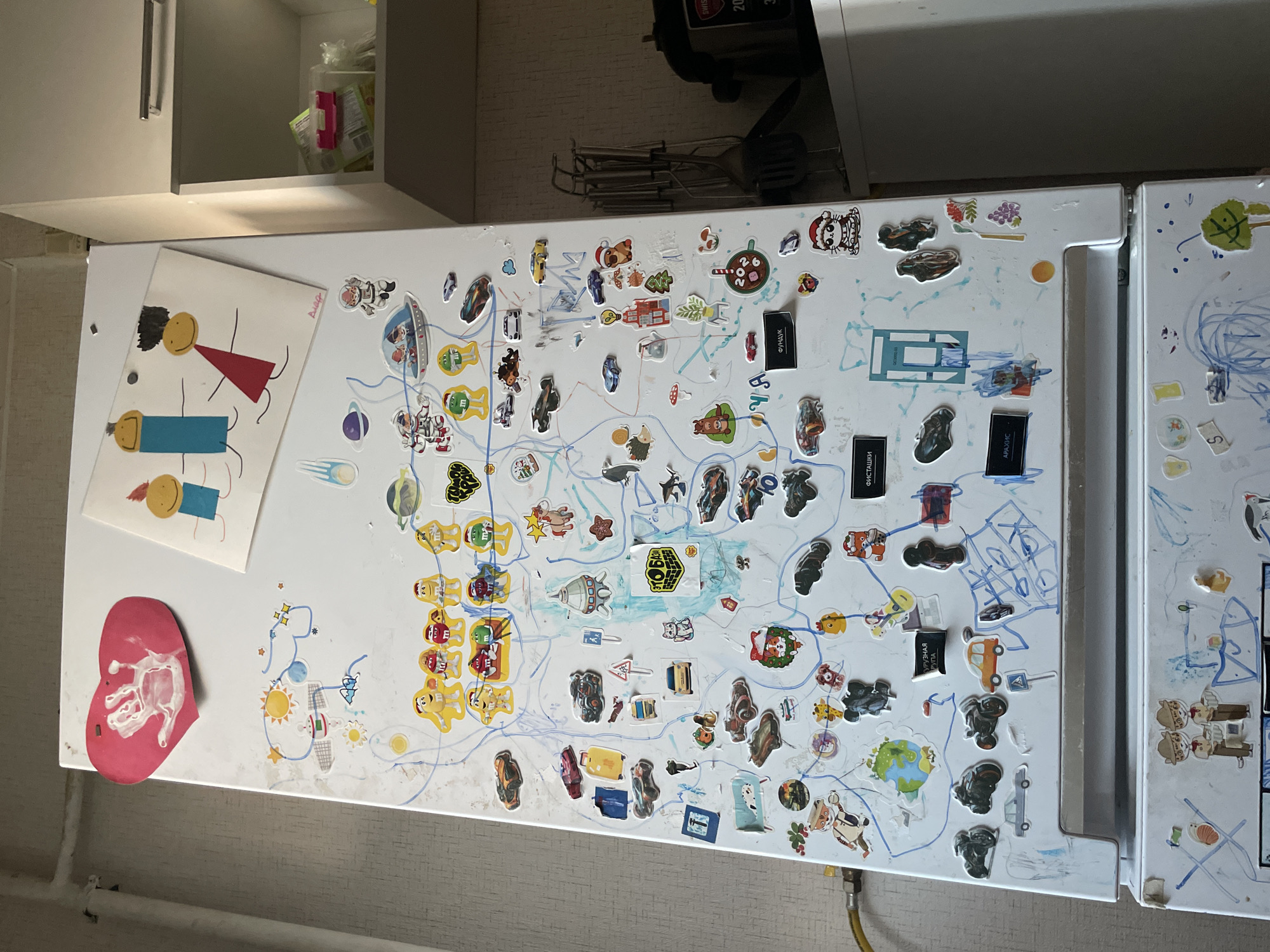The image shows a white refrigerator door covered in stickers and child-like drawings. The stickers depict various items such as cars, motorcycles, astronauts, animals, planets, and cartoon characters. There are also hand-drawn elements, including a red heart with a handprint, stick figures, and lines that appear to be part of a game or map. The refrigerator is in a domestic setting, with a wall visible in the background and part of a kitchen cabinet to the right. The overall impression is of a child's personalized and decorated appliance. The location provided, Laishevo, Russia, is noted but not visually verifiable from the image itself.